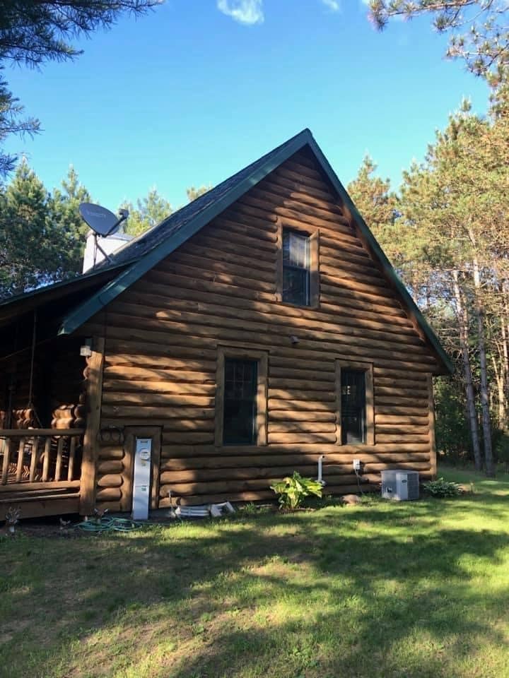 A rustic log cabin is set against a backdrop of tall pine trees. The exterior features a sloped roof with a chimney, multiple windows, and a porch area. The lush green lawn surrounds the cabin, enhancing the natural setting.
