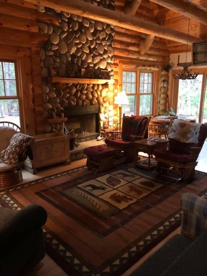 A spacious living area features a stone fireplace as the focal point, bordered by two rocking chairs with cushions. A wooden side table and a woven basket add warmth. Natural light filters through multiple windows, illuminating a patterned area rug depicting wildlife.