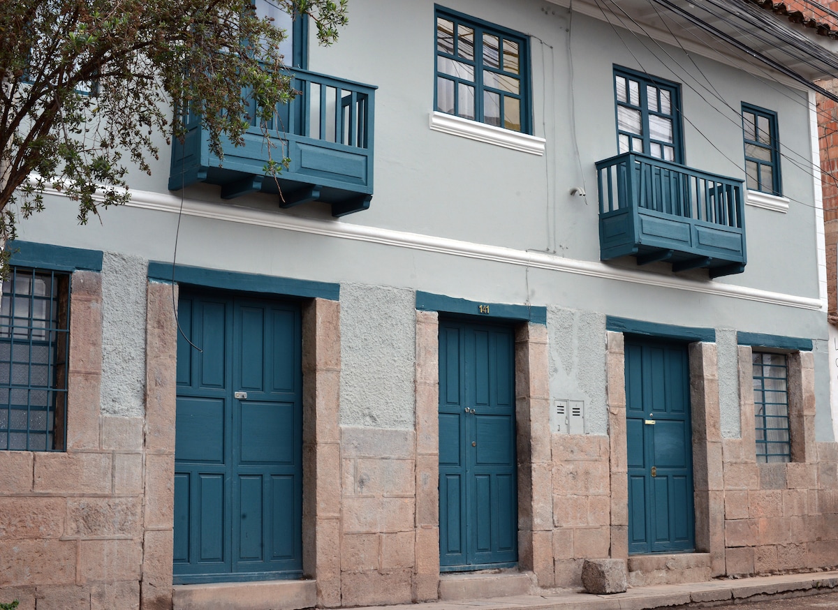 A colonial-style building is showcased, featuring a light blue facade accented by dark blue balconies. The sandy stone foundation highlights three symmetrical doors, framed by rectangular windows with black bars. The structure's charming architectural details reflect the traditional aesthetics of the neighborhood.