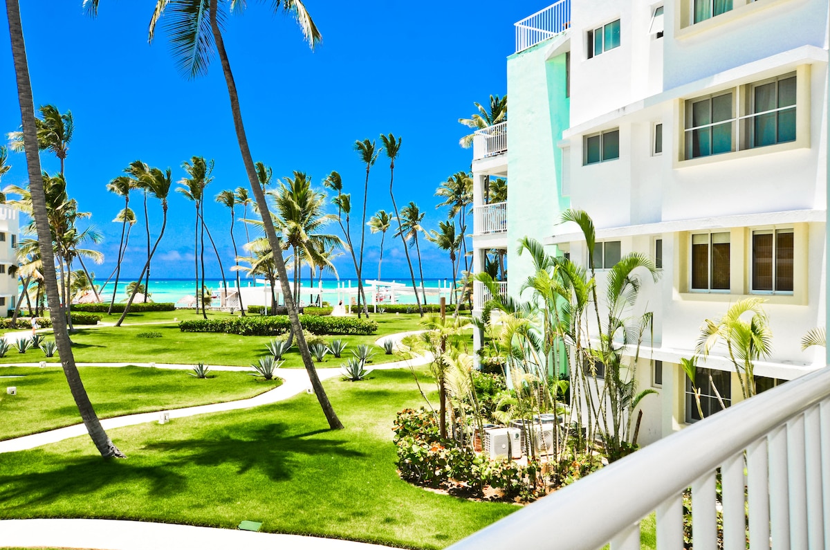A bright view from the balcony showcases lush green landscaping and vibrant palm trees swaying in the breeze. The turquoise ocean is visible in the background, enhancing the tropical ambiance of the surroundings. The building features large windows that establish a connection with the outdoor scenery.