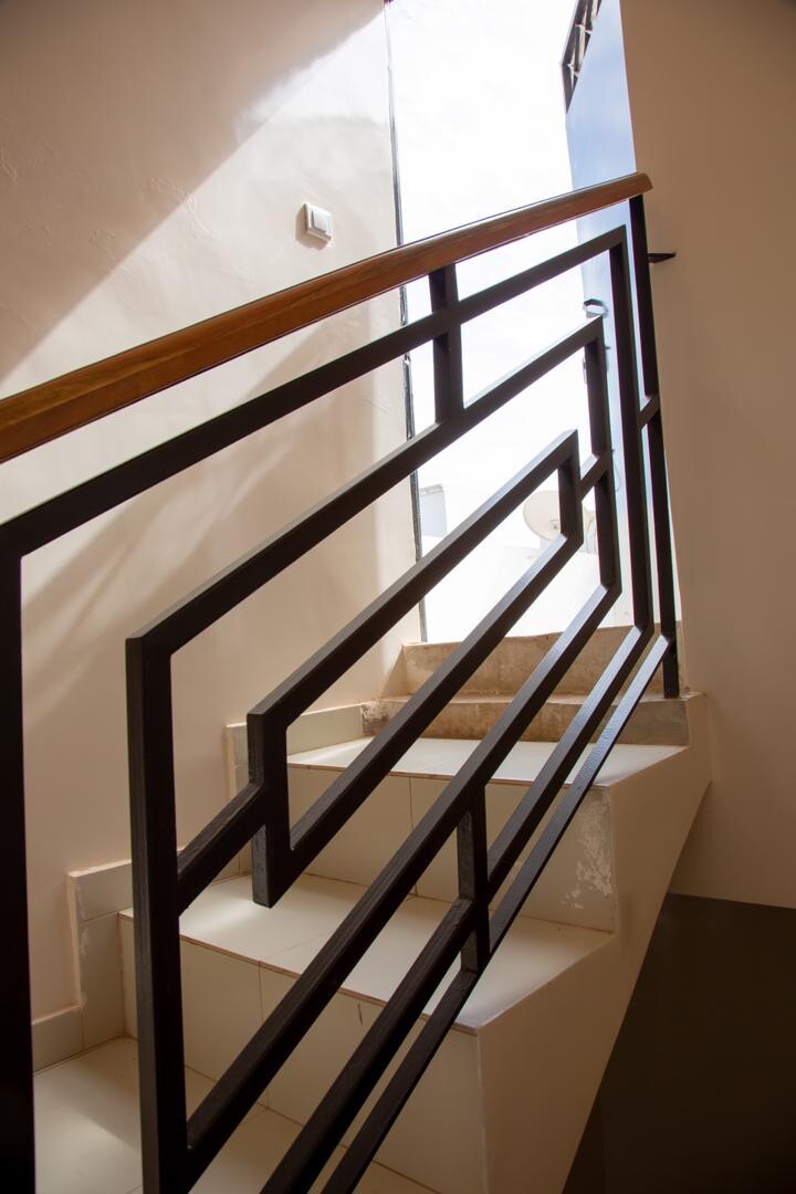 A well-lit staircase leads upward, featuring a sleek metal railing with geometric patterns. Natural light is allowed in through an open door at the top, illuminating the white walls and contrasting with the light-colored steps.