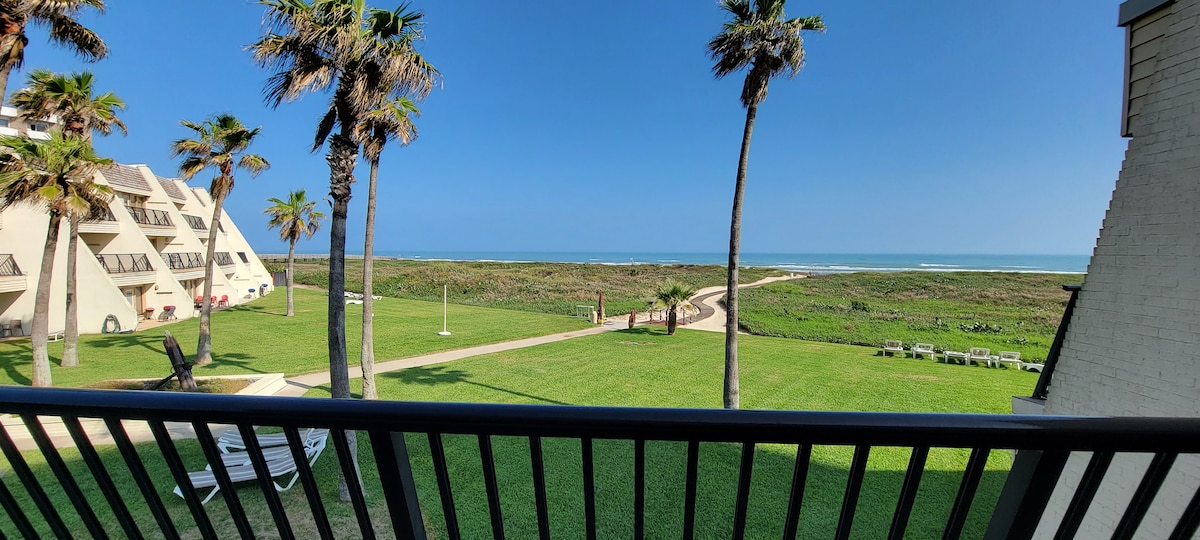 A lush landscaped area leads to the beach, framed by swaying palm trees. A clear blue sky meets the horizon, where the ocean waves gently break. An outdoor pathway is visible, inviting leisurely strolls along the beachfront.