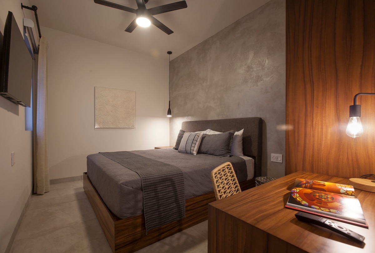 A modern bedroom features a spacious bed with gray bedding, complemented by a textured accent wall. A wooden bedside table holds magazines, while a round, woven chair adds a subtle design element. Soft lighting from pendant fixtures enhances the serene ambiance.