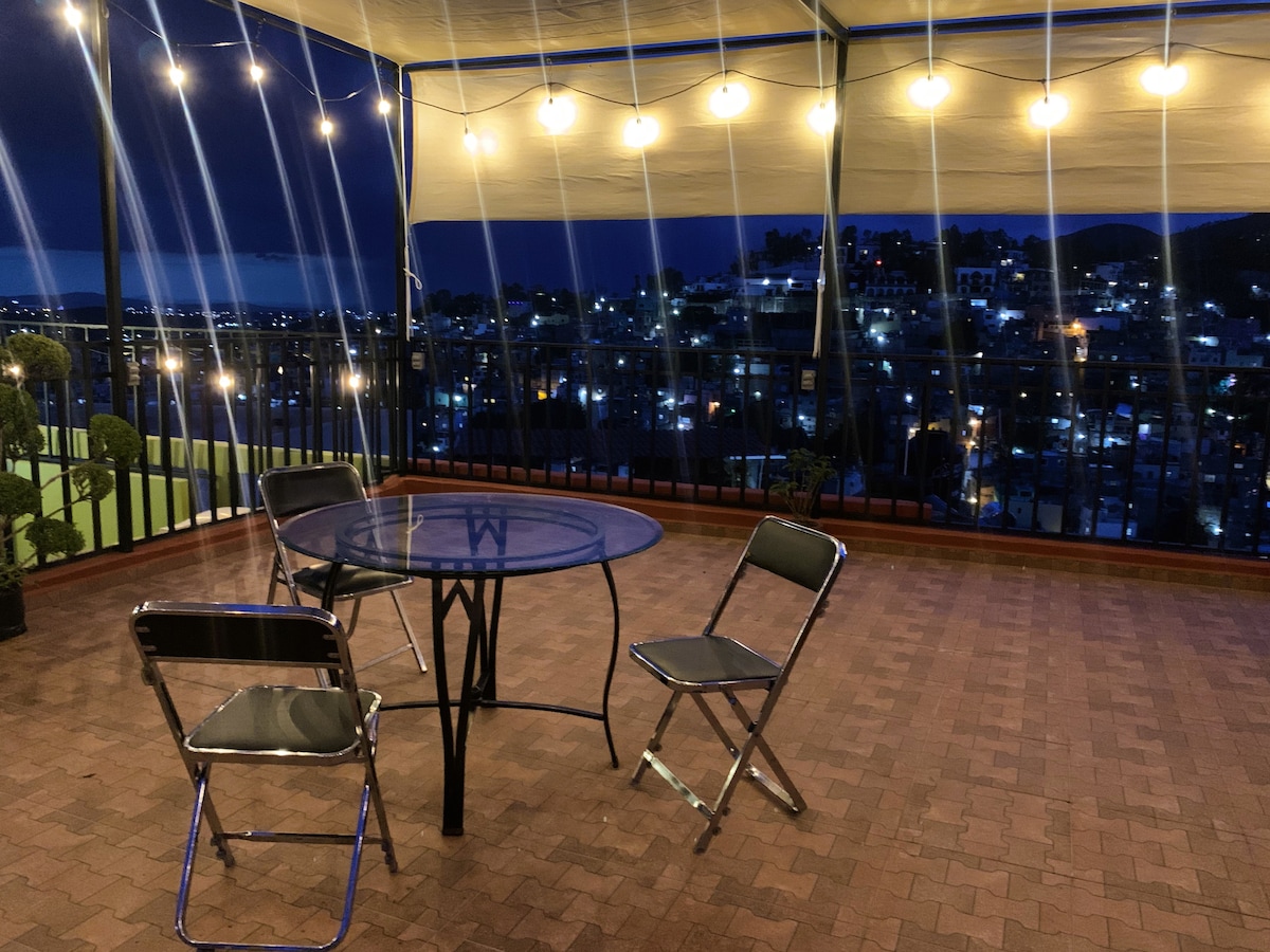 An outdoor terrace is equipped with a round table and four chairs, offering views of the city illuminated at night. Soft overhead lighting adds to the ambiance, creating a welcoming environment. Potted plants are positioned around the space, enhancing its design.