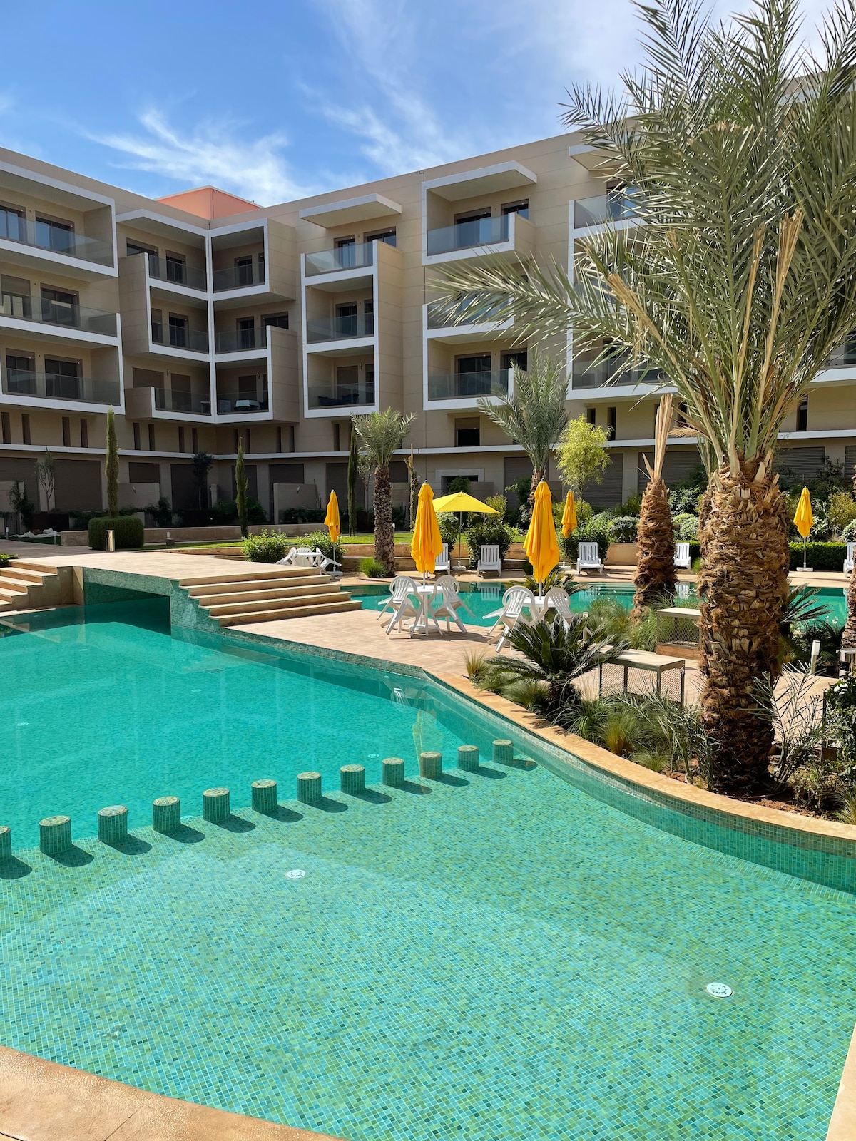 A modern residential complex features an outdoor pool surrounded by palm trees and lounge chairs with yellow umbrellas. The pool's vibrant blue water reflects the sky, while landscaped greenery adds a refreshing touch.