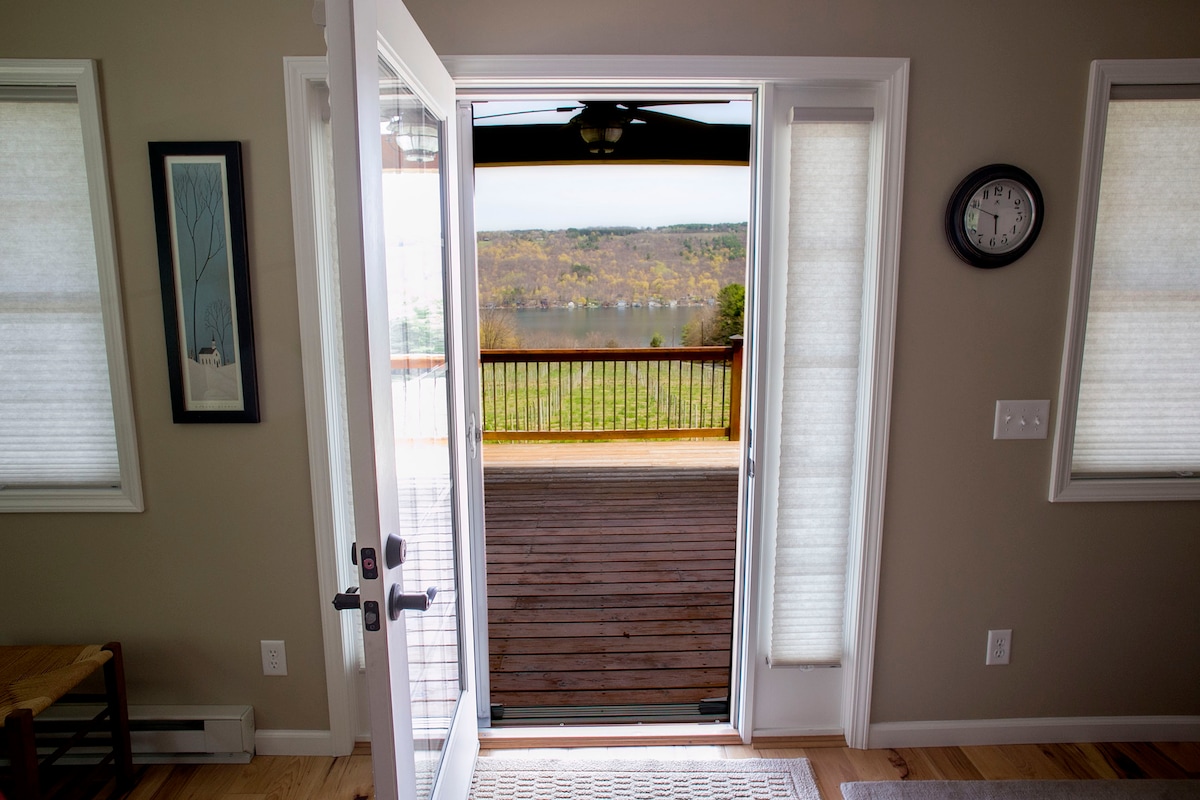 An open door reveals a view of the expansive deck and surrounding landscape, leading to Keuka Lake. Light-filtering shades adorn the windows, enhancing the ambiance. A wall clock and framed artwork are visible, contributing to the space's design.