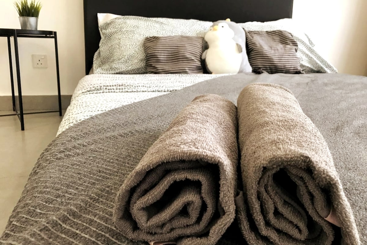 A spacious bed is neatly arranged with patterned bedding and decorative pillows. Two rolled towels are positioned at the foot of the bed. A plush toy sits on the bed, and a small table with a plant can be seen beside it.