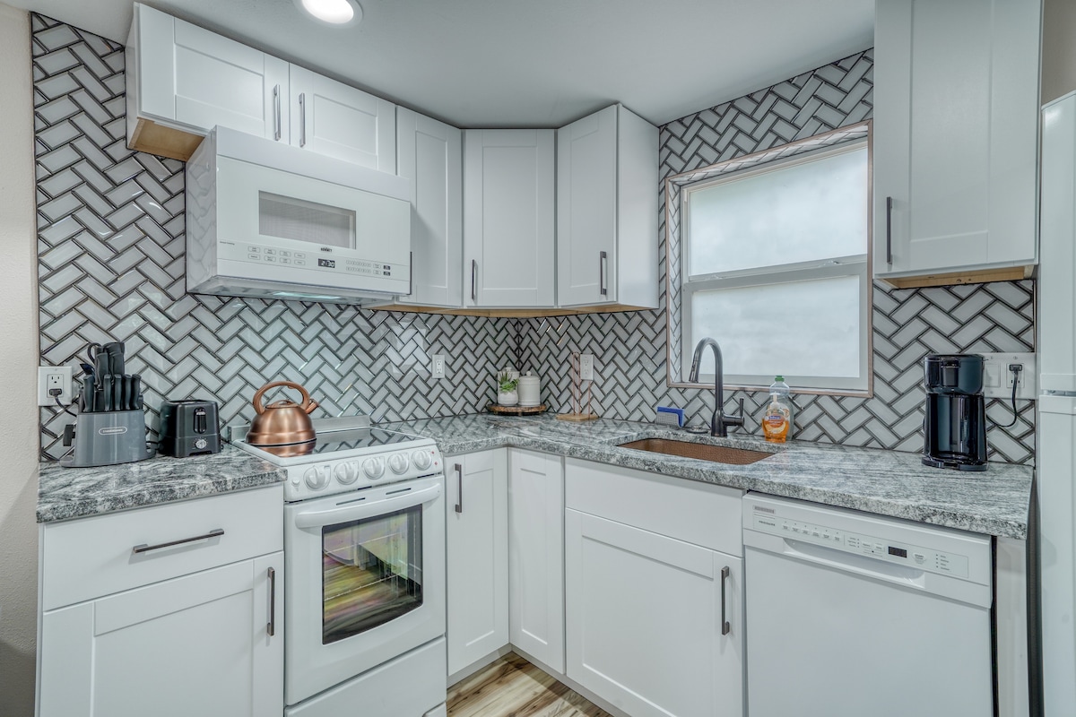 The kitchen features modern cabinetry and a stylish backsplash with a herringbone pattern. Essential appliances include a gas stove, microwave, dishwasher, and coffee maker. Ample counter space is provided along with a stainless steel sink overlooking a window, allowing natural light into the area.