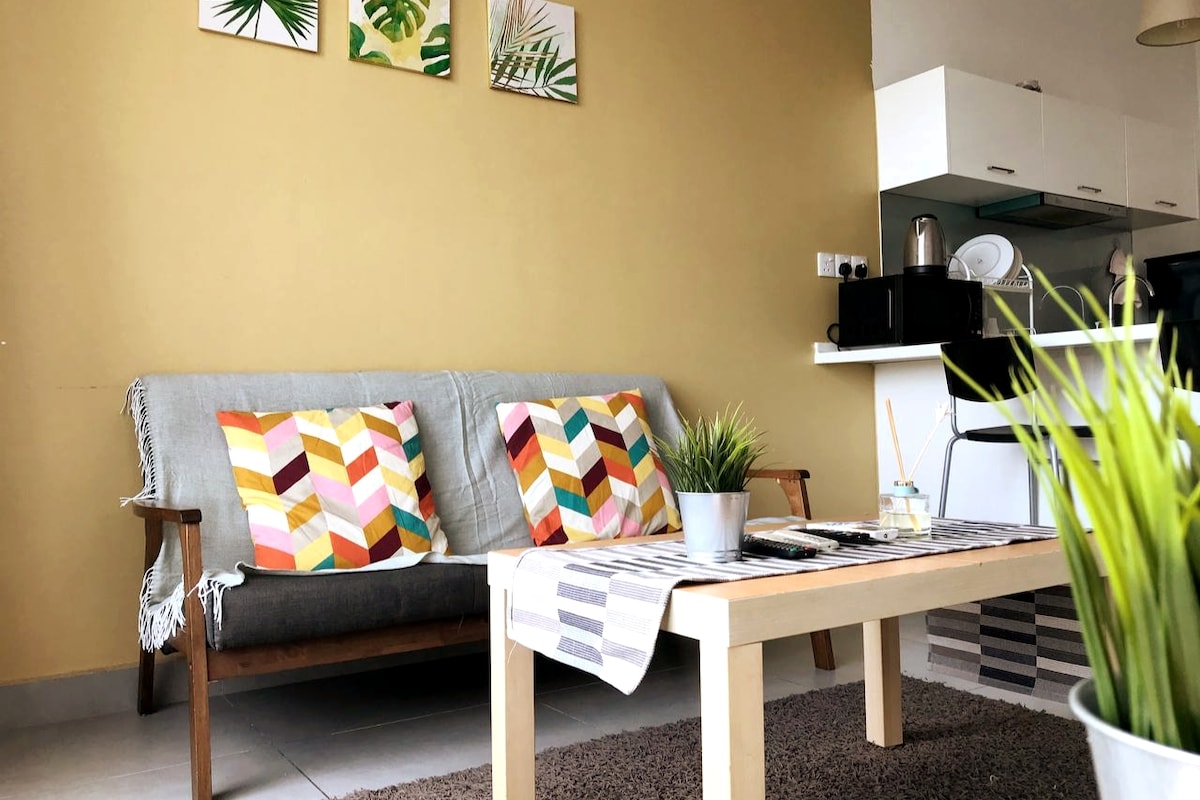 A cozy seating area is presented, featuring a light grey fabric sofa adorned with colorful geometric-patterned cushions. A small table is positioned in front, complemented by a potted plant. In the background, a compact kitchen with modern appliances is visible.