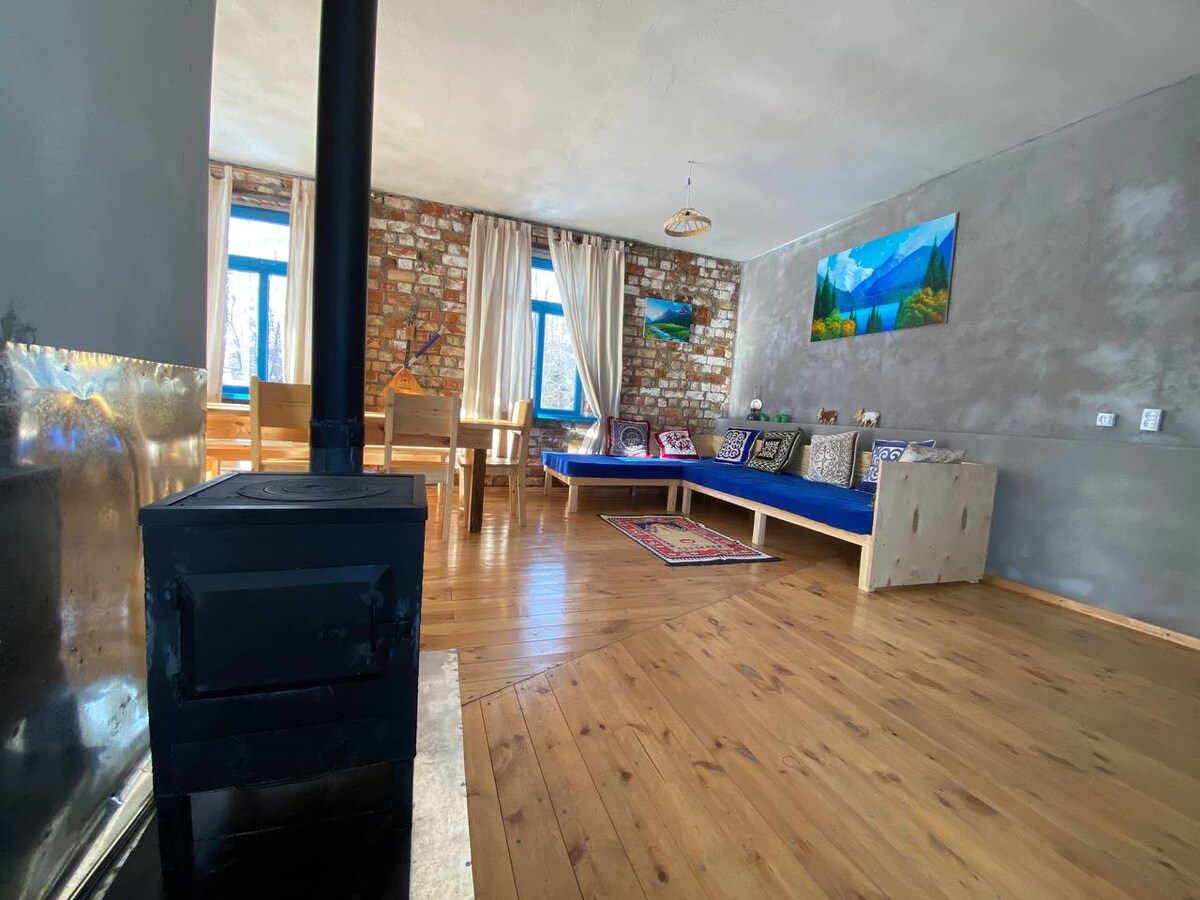 A spacious living area features a cozy seating arrangement with blue cushions along the wall. A rustic wooden table is surrounded by chairs, while large windows invite natural light. A small wood stove adds warmth, against a backdrop of exposed brick and grey walls.