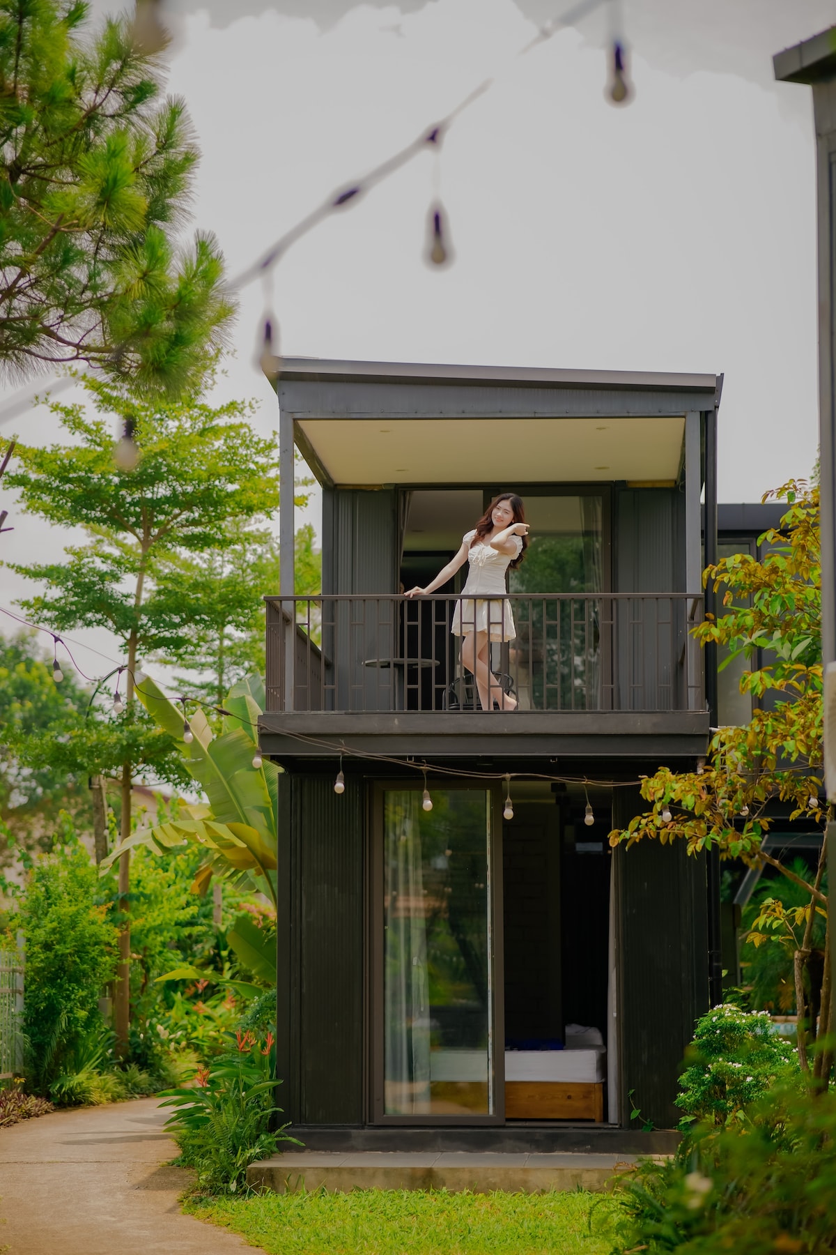 A modern two-story structure is showcased, featuring a balcony with a person standing gracefully. Surrounding greenery includes tall trees and flowering plants, while soft lighting is provided by decorative string lights. Large glass doors reveal a glimpse of the indoor space.