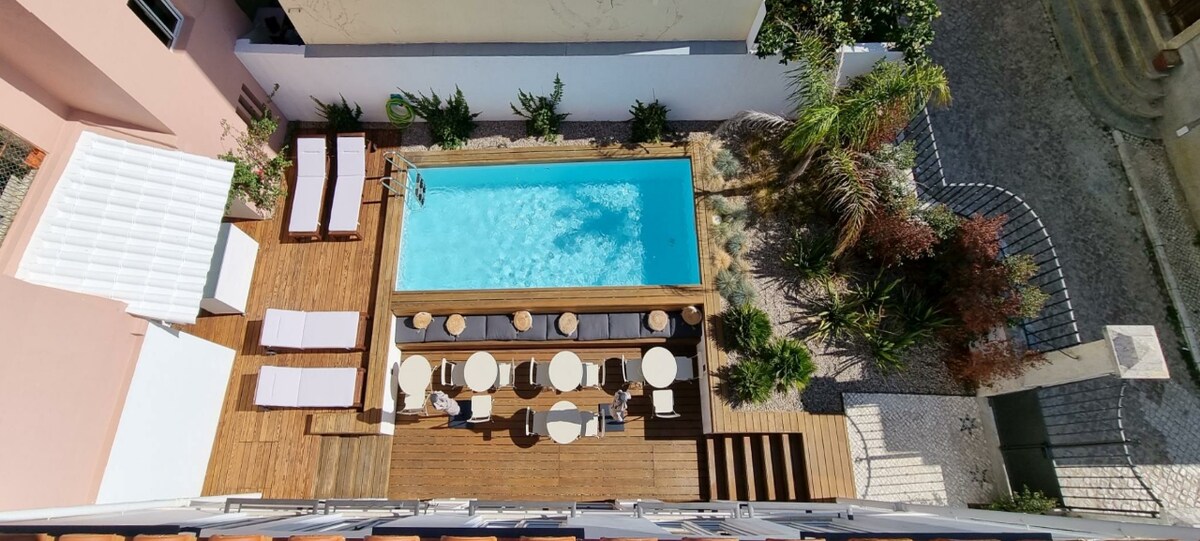 An aerial view reveals a swimming pool surrounded by wooden decking. Lounge chairs and elegant tables offer space for relaxation. Lush greenery provides a natural border, enhancing the outdoor setting, while the layout encourages social gatherings.