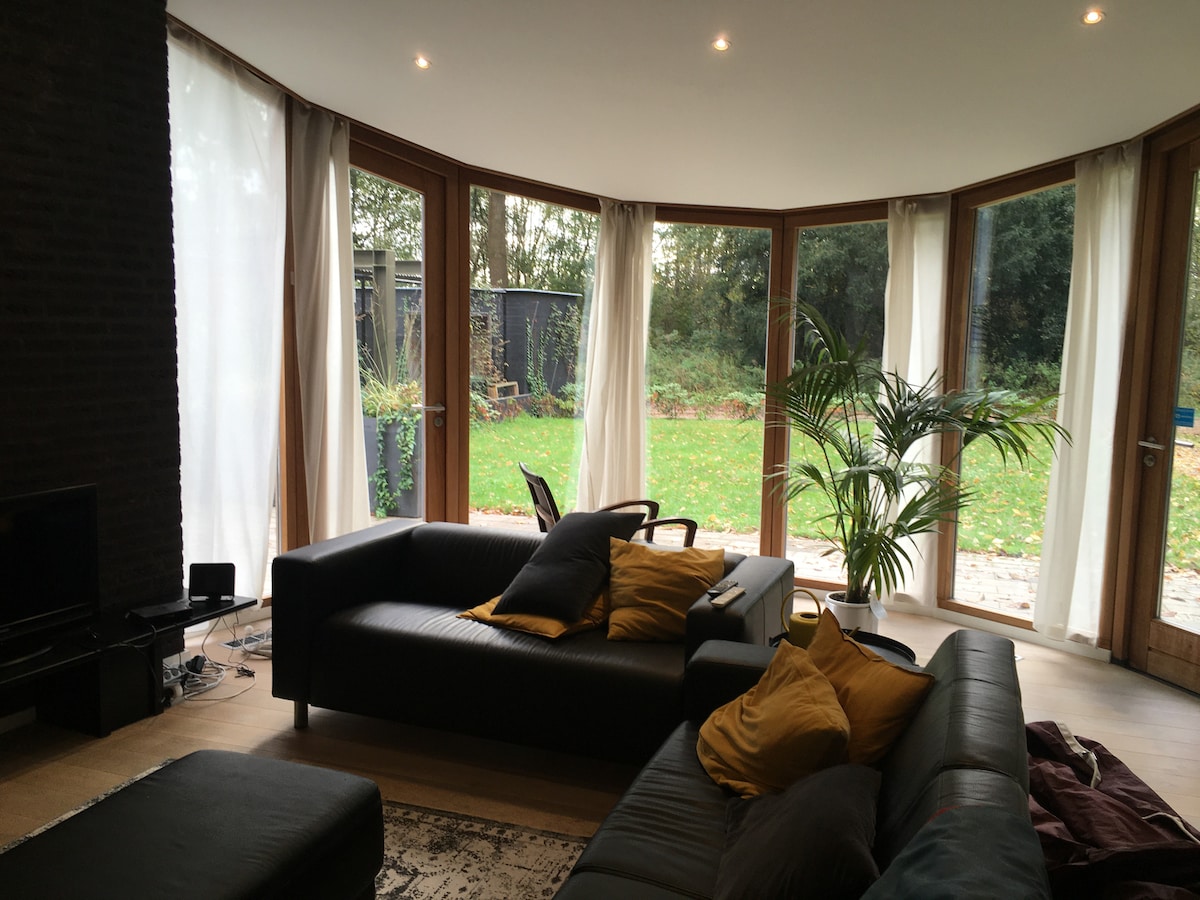 A spacious living area features large curved windows that invite natural light and offer views of the surrounding greenery. Two comfortable sofas adorned with throw pillows are positioned for relaxation. A plant adds a touch of nature to the modern decor.