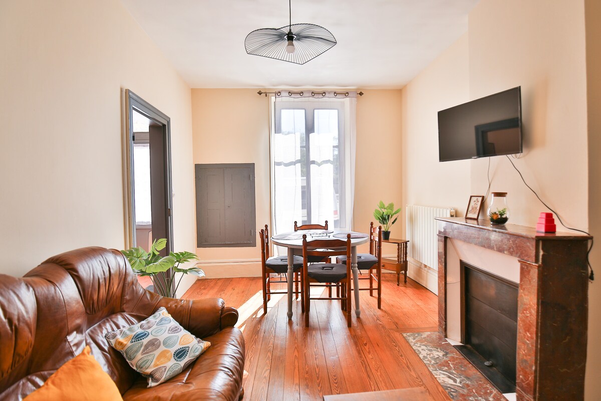 A living area features a comfortable brown leather sofa adorned with colorful cushions. A wooden dining table with four chairs is positioned nearby, accompanied by a plant. Natural light streams through a large window with light curtains, complementing the warm wooden flooring.