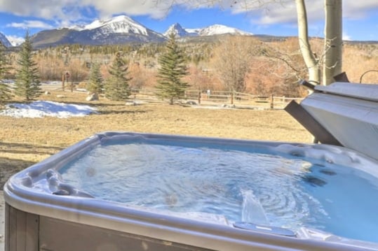 A private hot tub is positioned outdoors, surrounded by a serene landscape featuring snow-capped mountains in the distance. The area is complemented by scattered trees and open space, providing a tranquil setting for relaxation.