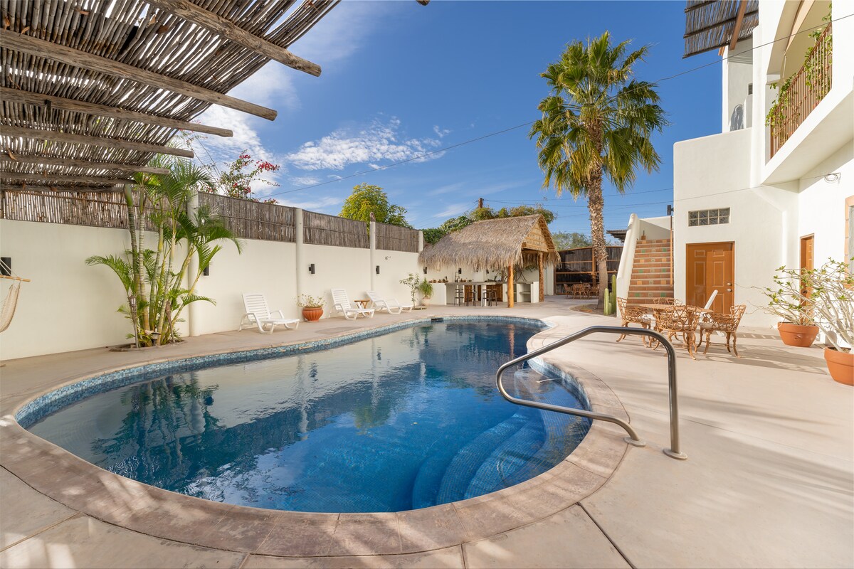 A serene pool area is surrounded by lush greenery and palm trees. A spacious pool with inviting blue water reflects the sky above. A palapa-style outdoor kitchen and bar are visible in the background, providing shaded seating options beside the pool.