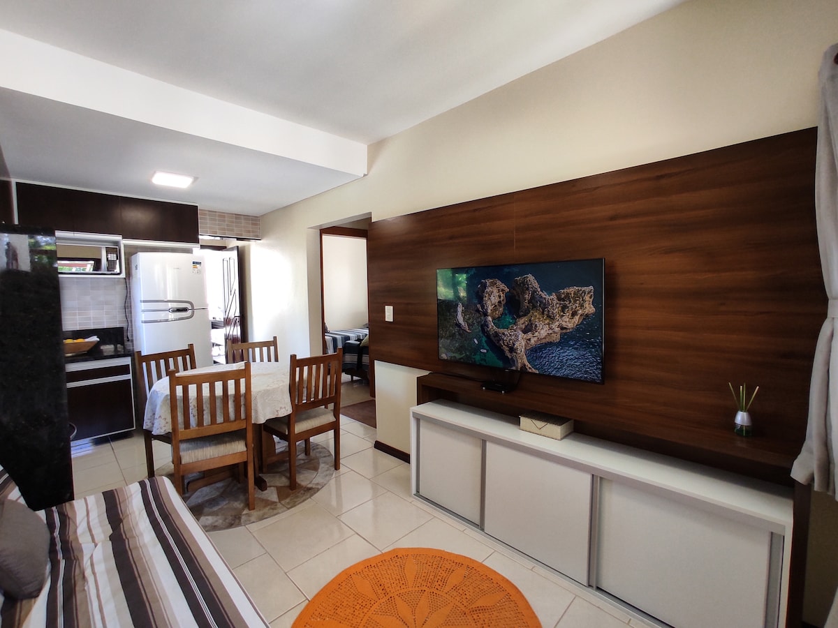 A living area features a modern wooden panel with a flat-screen TV mounted above a sleek white cabinet. A round orange rug sits beneath a wooden dining table surrounded by four chairs, while the kitchen area and another room are visible in the background.