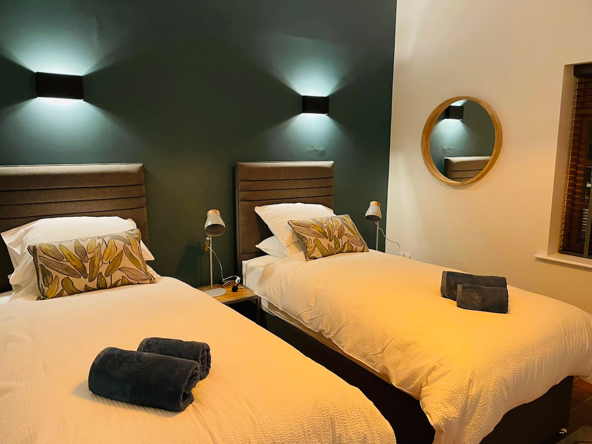 The bedroom features two spacious beds with crisp white bedding, complemented by decorative pillows. Soft lighting is provided by wall sconces above each bed, while a round mirror adds a touch of elegance to the wall. Fresh towels are neatly placed at the foot of each bed.