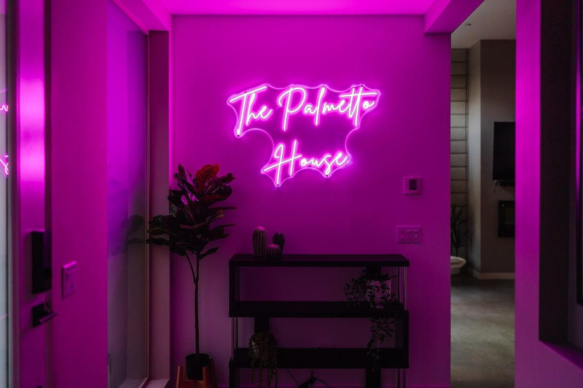 A modern entryway is illuminated by a pink neon sign displaying 'The Palmetto House.' A decorative plant is positioned next to a sleek console table, creating a welcoming ambiance as natural light enters from adjacent areas.