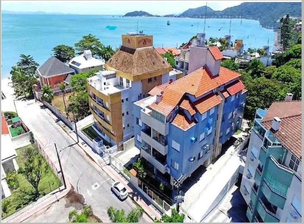 An aerial view captures a cluster of residential buildings with varied architectural styles along a coastal road. The blue waters and distant hills are visible, providing a scenic backdrop. The streets are tree-lined, suggesting a relaxed neighborhood atmosphere near the beach.