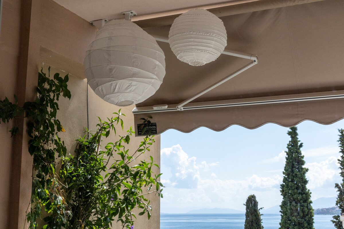 Outdoor space features hanging white lanterns, providing soft illumination. A canopy shades the area, while lush greenery adds a natural element. The view of the sea and distant hills can be seen through the open space, enhancing the tranquil atmosphere.