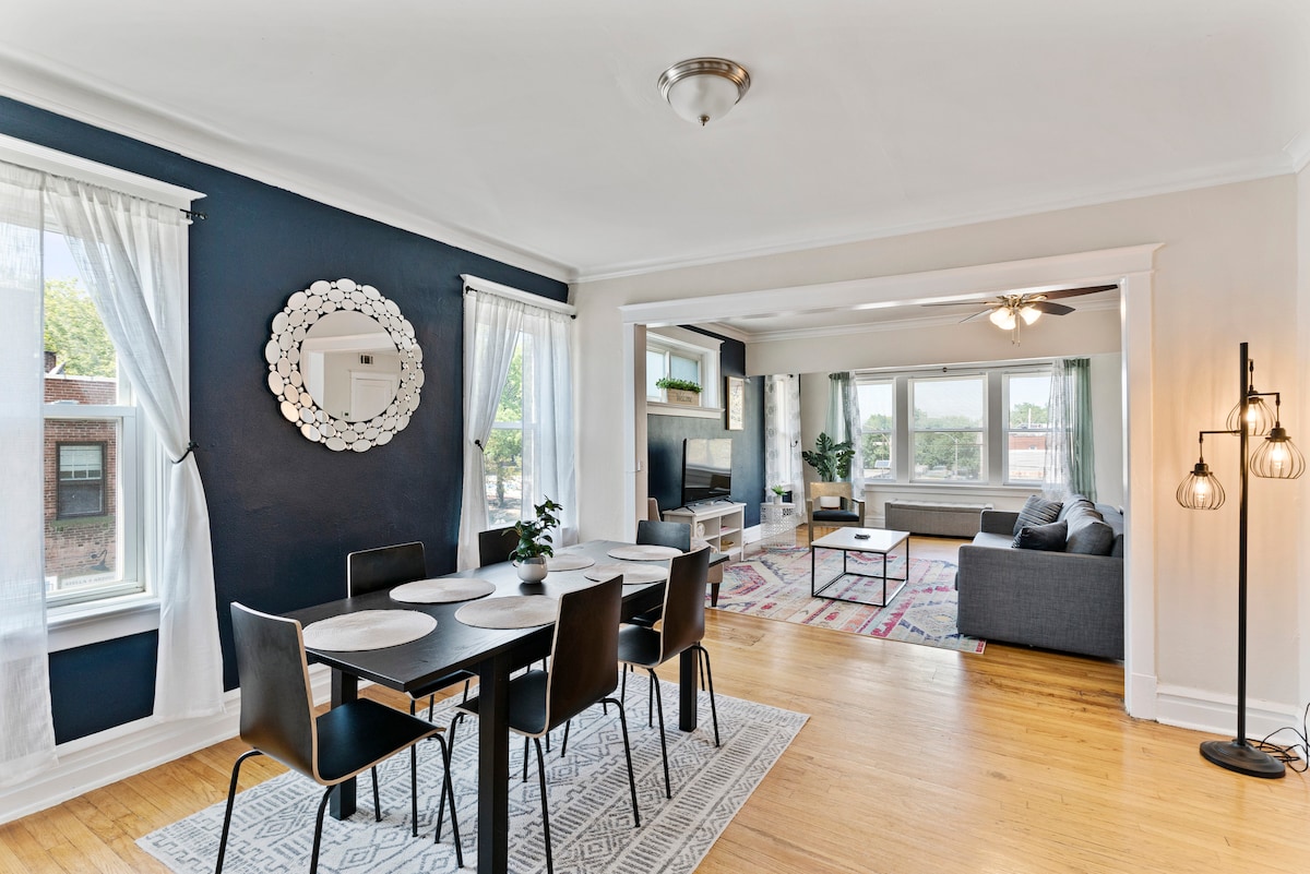 An open concept living and dining area showcases hardwood floors and neutral wall tones. A dining table with four chairs is positioned next to large windows, while a cozy seating arrangement with a gray sofa is visible in the background. A decorative mirror enhances the space.