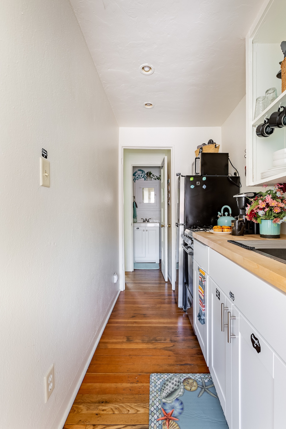 A narrow kitchen corridor is displayed, featuring wooden flooring that extends throughout. Cabinets and appliances in white and black are visible, with a sink and countertop neatly arranged. A door at the end leads to a cozy bathroom, while decorative items and flowers add a touch of charm.