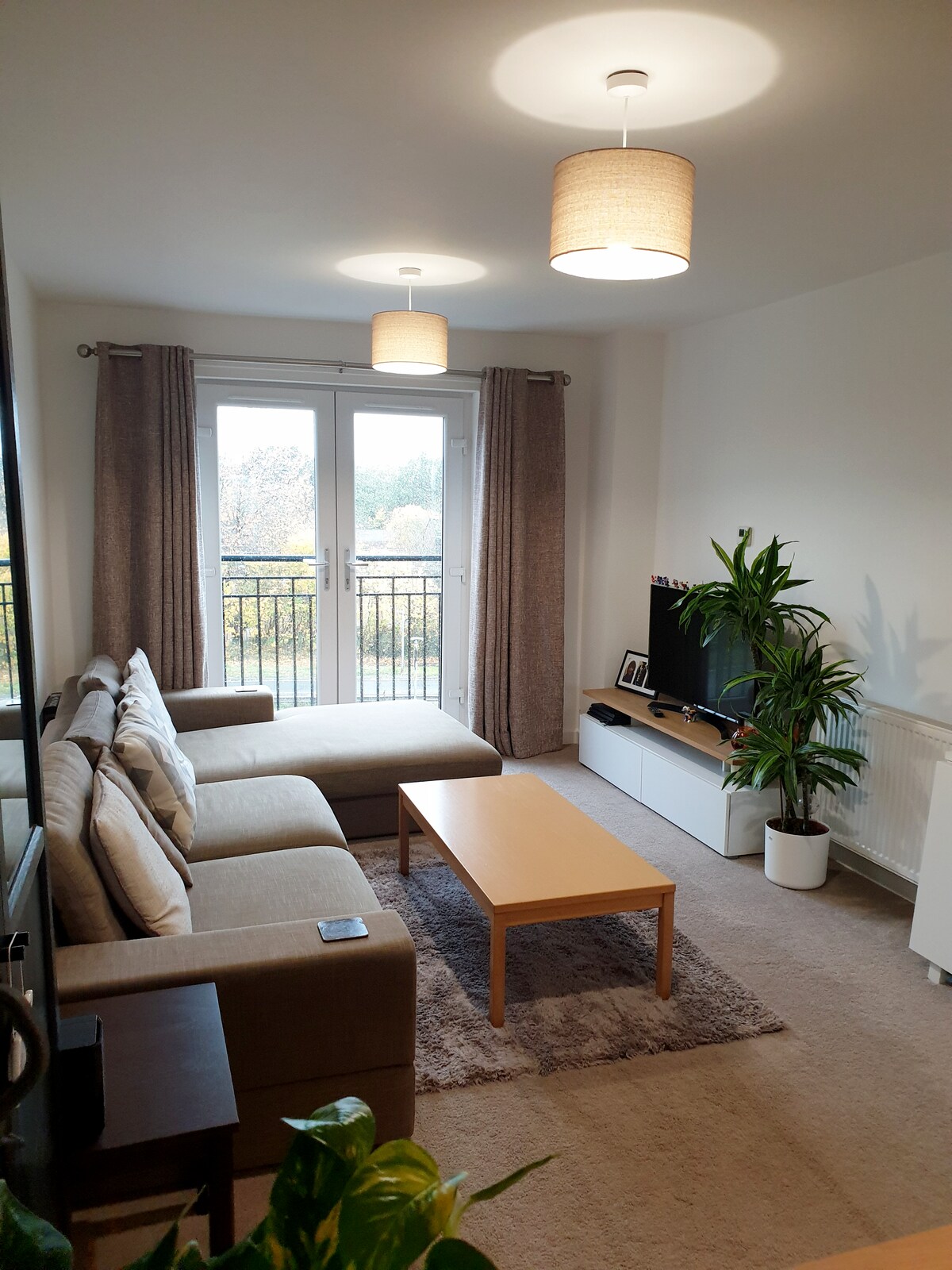 A bright living area features a beige sofa positioned near a wooden coffee table. Natural light floods in through the balcony doors, which lead to a small outdoor space. A green plant adds a touch of nature, while a flat-screen TV sits on a white media unit.
