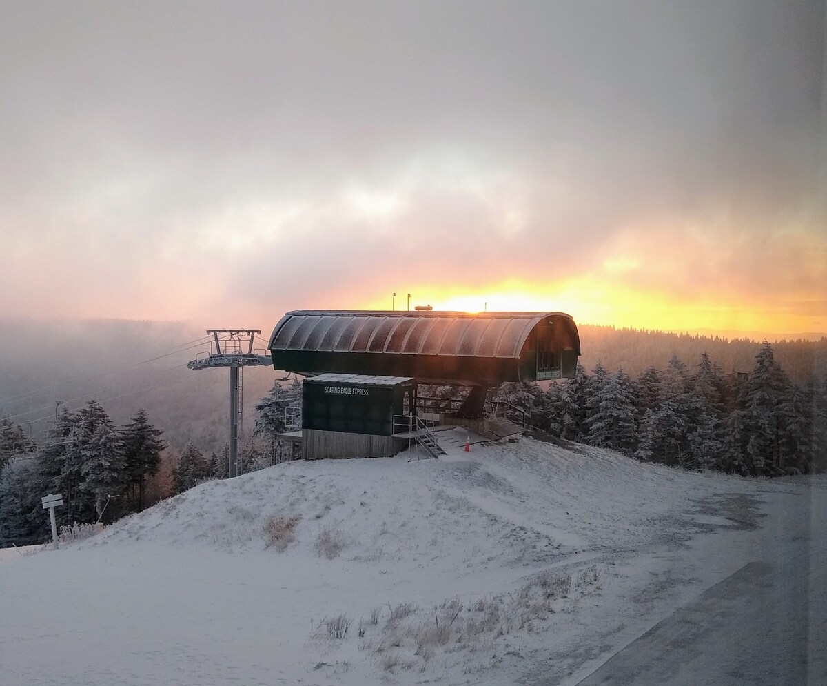 A ski lift station is viewed against a backdrop of a winter sunset, with hues of orange and pink illuminating the sky. Snow-covered slopes extend below, and trees are blanketed in snow, creating a serene mountain landscape.