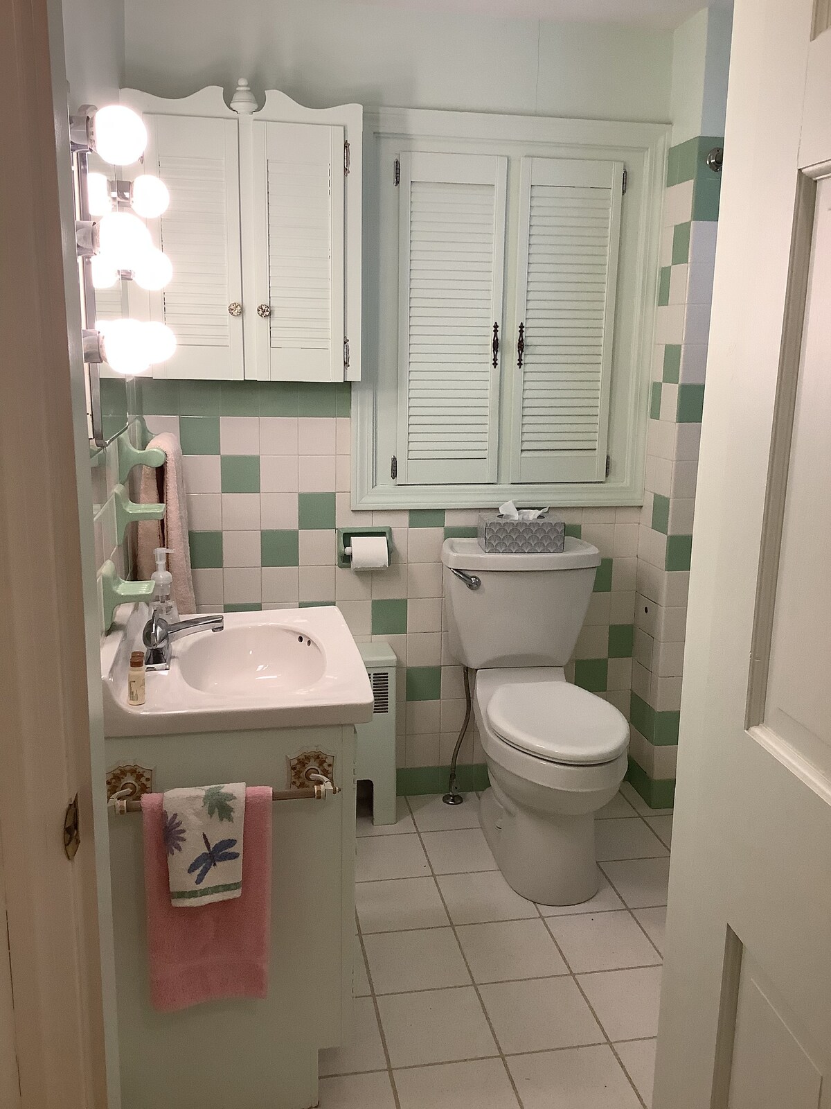 A compact bathroom features pastel green and white tiles. A single sink with a mirrored cabinet above is located beside a toilet. Soft lighting is provided by wall-mounted fixtures. Fresh towels are neatly displayed, and a decorative storage box sits on the counter.