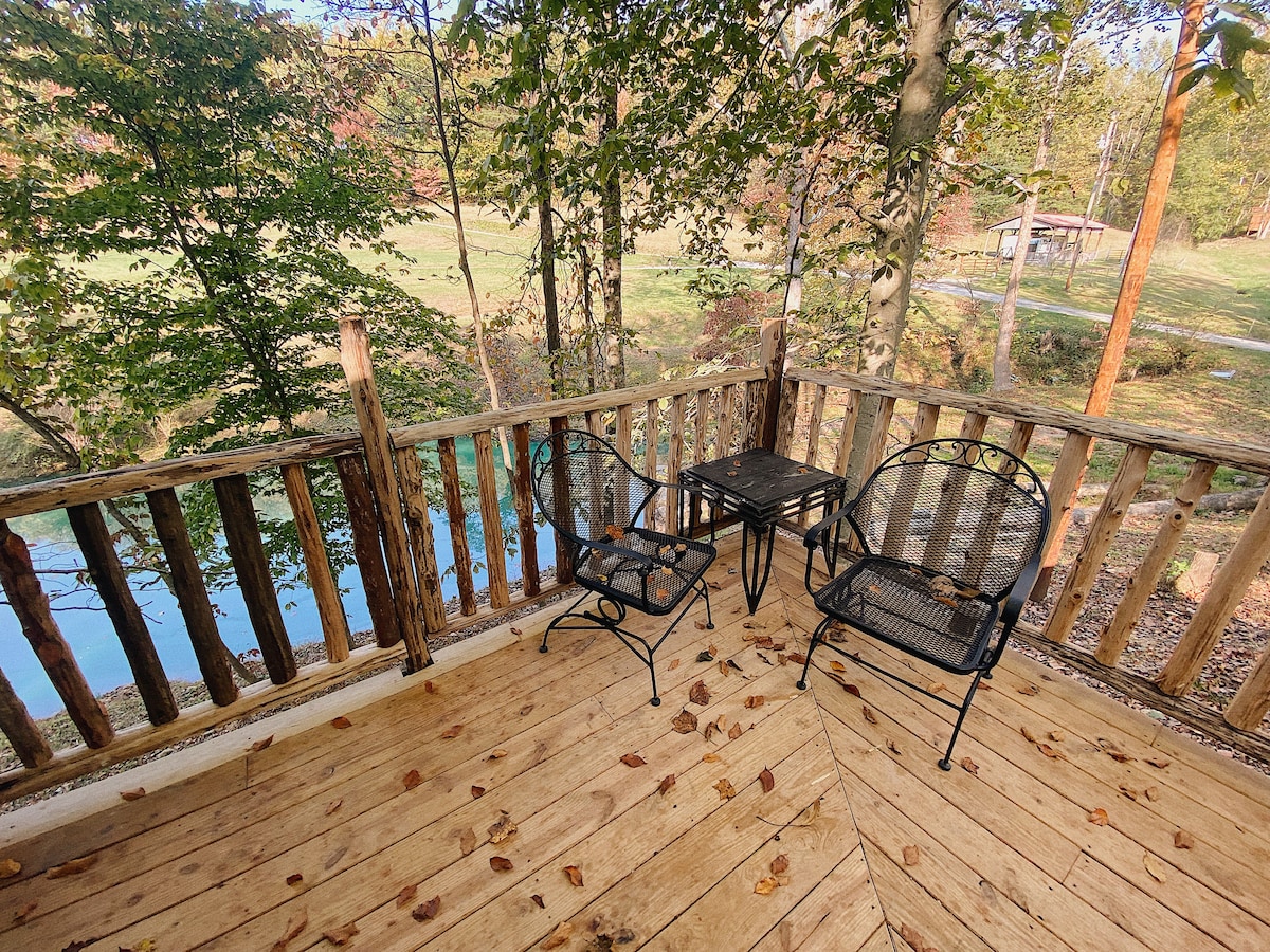 A serene outdoor seating area is featured, consisting of two metal chairs and a small table on a wooden deck. Surrounding trees provide a natural backdrop, while fallen leaves add a touch of seasonal charm. A glimpse of water can be seen through the foliage.