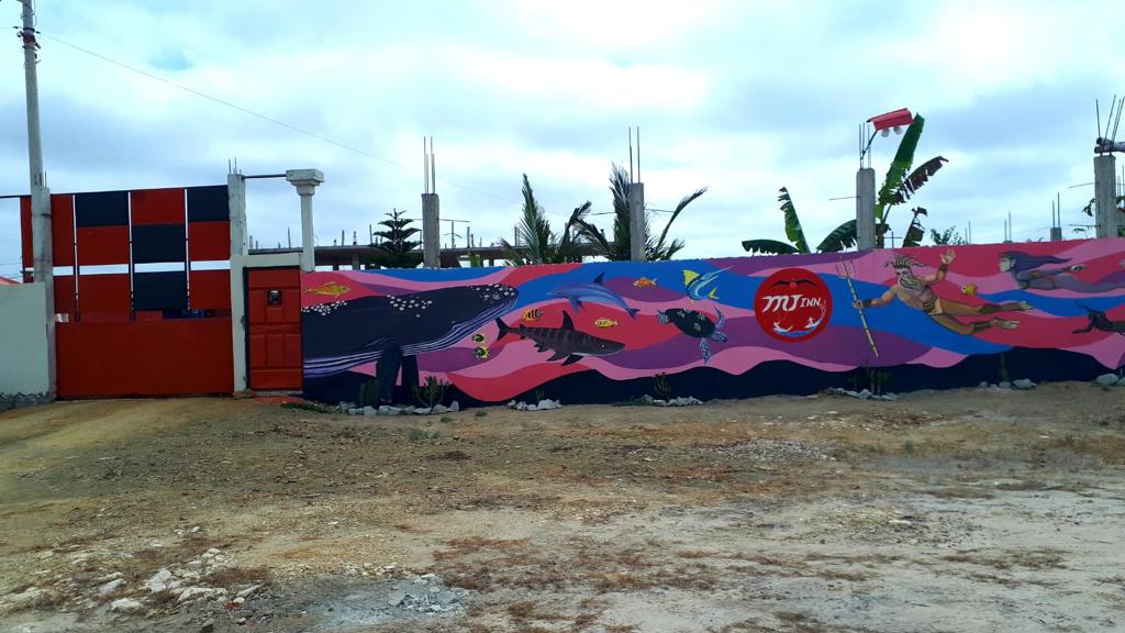 An eye-catching mural spans the wall, depicting vibrant aquatic life against a pink and blue background. The entrance features a bold red gate, flanked by green banana plants and a clear sky, contributing to the welcoming exterior of the property.
