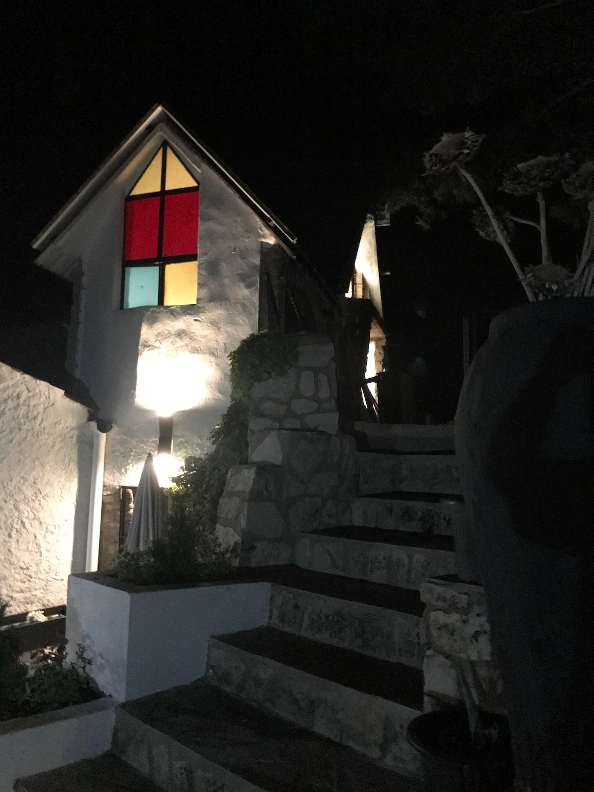 A charming exterior view of a white building at night, featuring a colorful stained-glass window that includes red and yellow panes. A stone staircase leads up to the entrance, illuminated by soft outdoor lighting, enhancing the inviting atmosphere of the property.