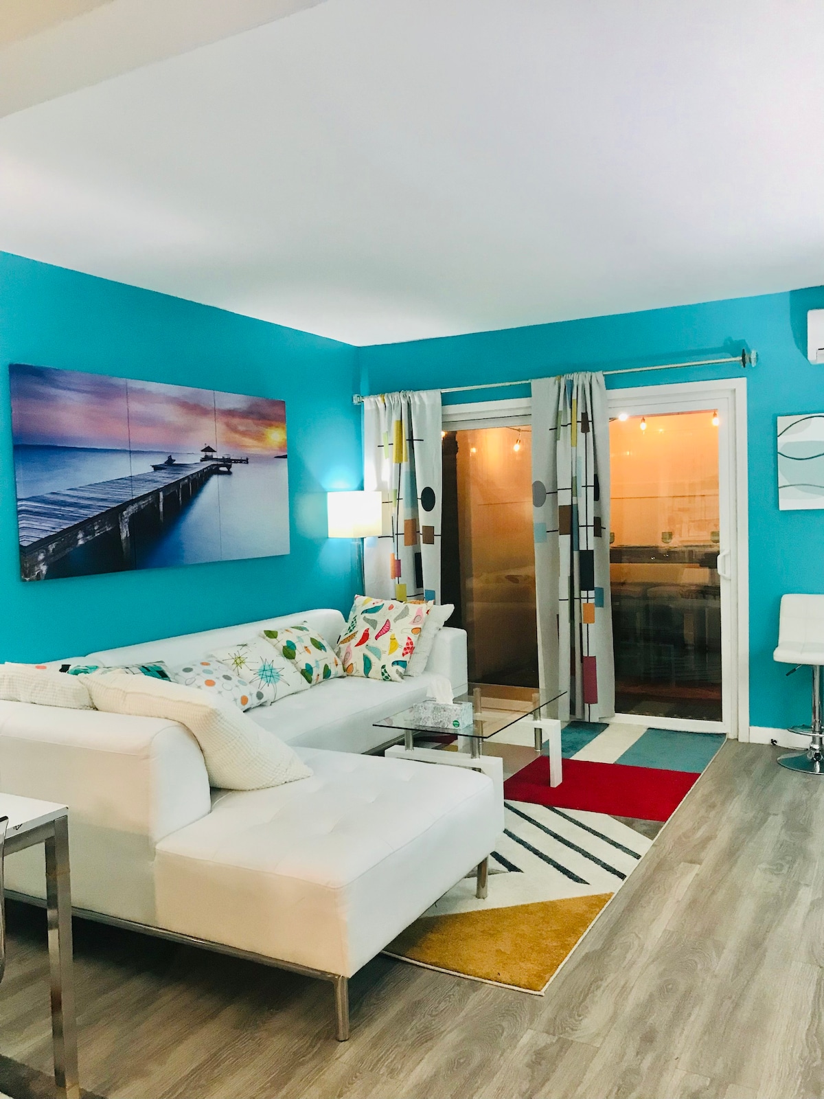 A brightly colored living space features a white sectional sofa adorned with patterned cushions. A glass coffee table sits on a multi-colored area rug. Large glass doors lead to a patio, while a wall art piece depicting a pier adds interest to the turquoise walls.