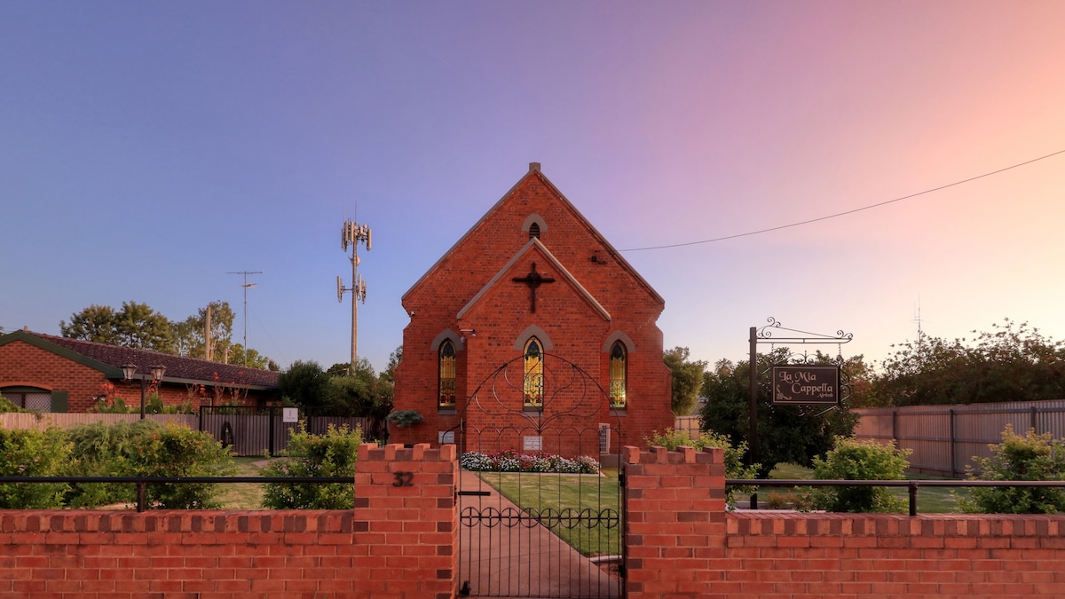 The exterior of La Mia Cappella is captured at twilight, featuring a historic brick facade adorned with a prominent stained glass window. The property is framed by hedges and decorative plantings, with a gated entrance leading to the church and a sign indicating its name.