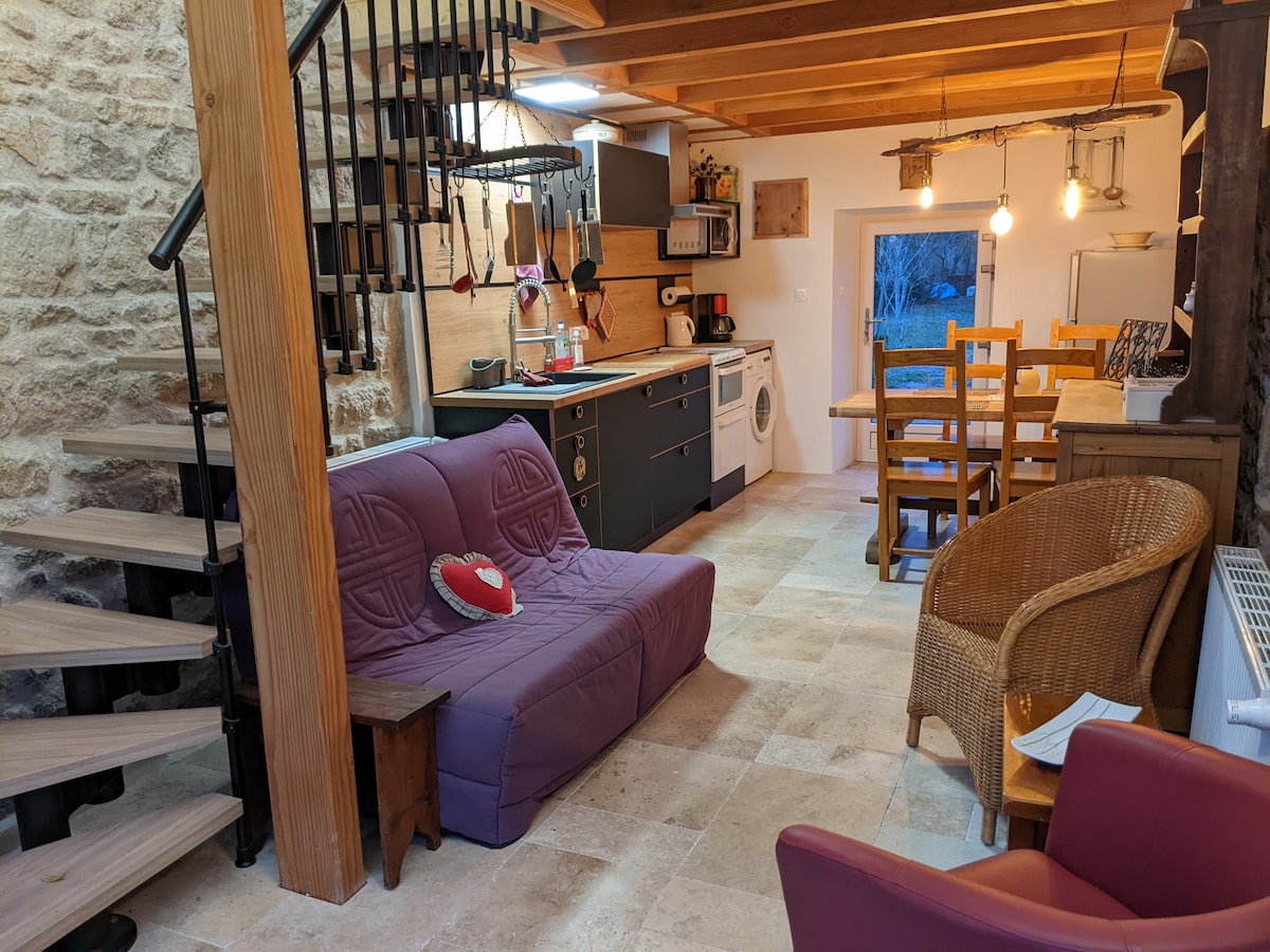 A cozy living area features a purple sofa, a wooden dining table, and chairs. A spiral staircase leads to a higher level, while a fully equipped kitchen is visible. Soft lighting from overhead fixtures creates a welcoming ambiance, complemented by natural light from the large window.