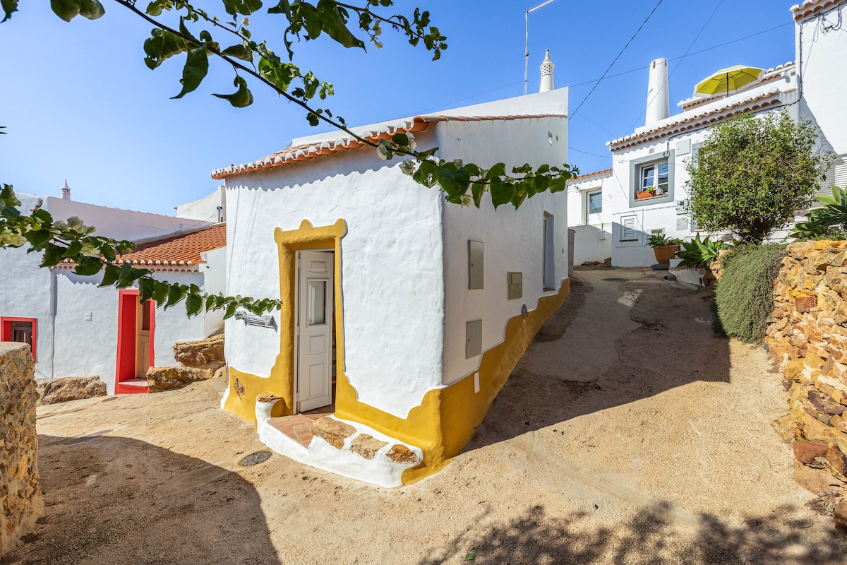 A charming white house with yellow accents is situated on a quiet, sloped street in a historic village. The surrounding area features natural rocky elements and greenery. The structure has a simple door and small windows that complement the rustic style of the neighborhood.