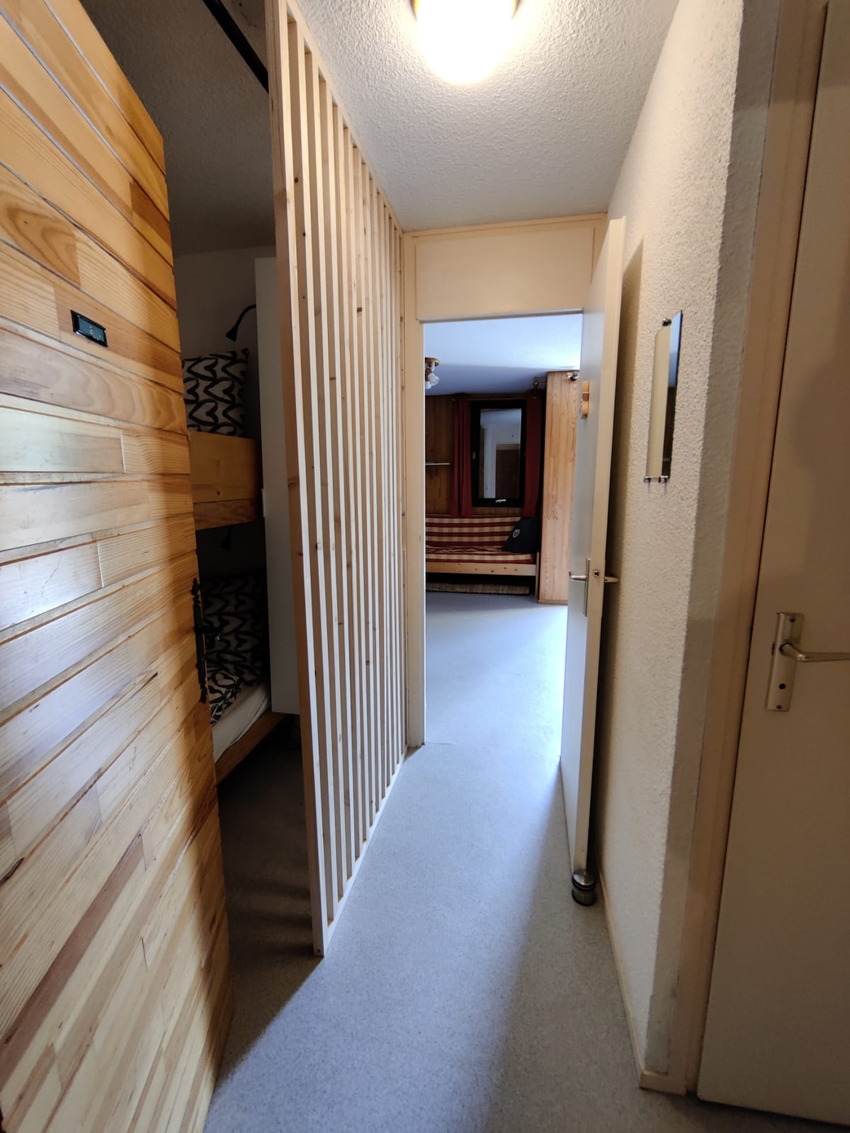 A hallway is visible, featuring wooden paneling on one side and a doorway leading to a cozy sleeping area. Soft carpet flooring extends throughout the space, and light illuminates the corridor from overhead lighting.