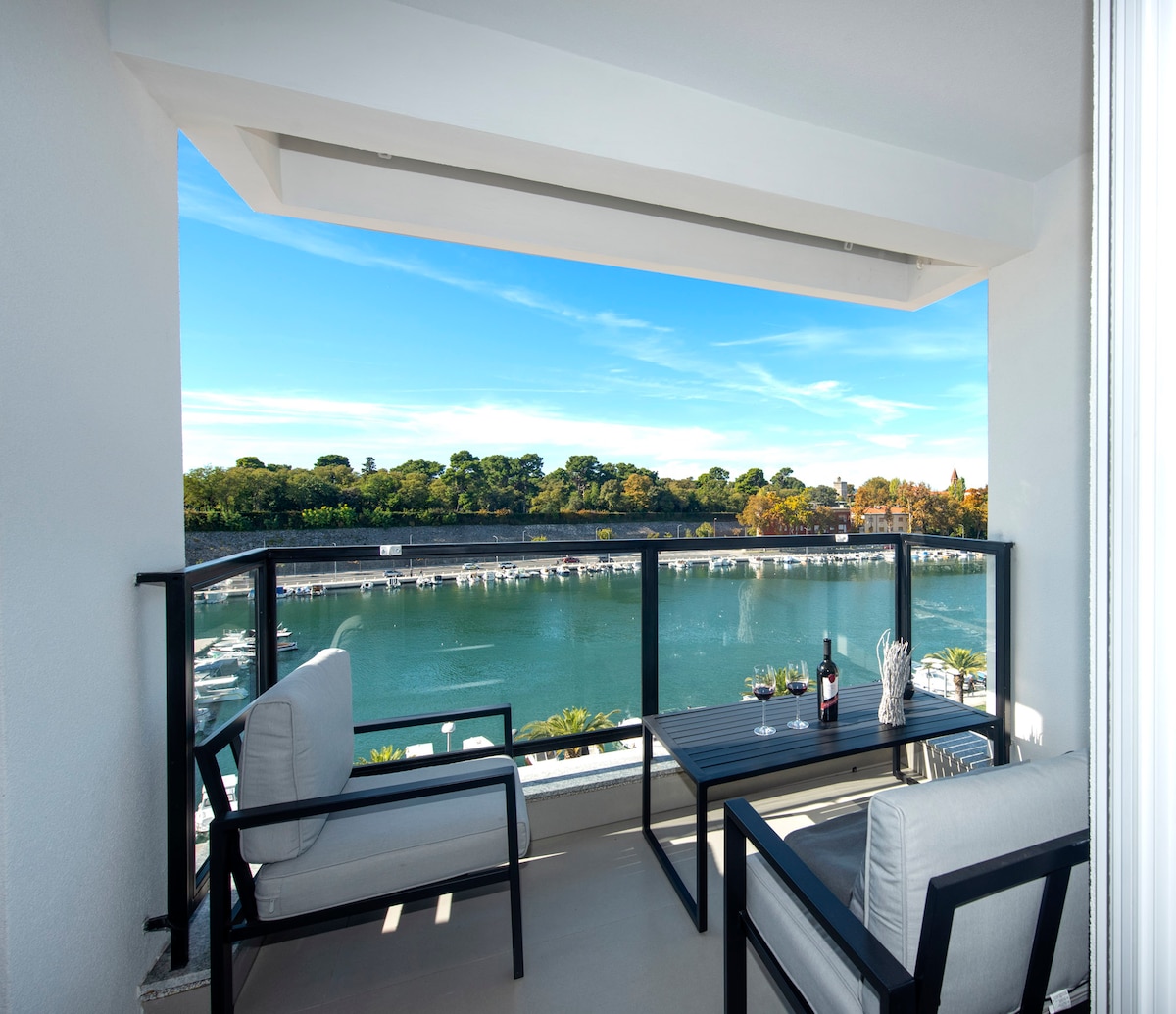 An outdoor balcony features a table with two chairs, overlooking the water. The scene displays lush greenery and distant trees under a clear blue sky. A bottle and glasses are positioned on the table, providing a serene spot for relaxation.