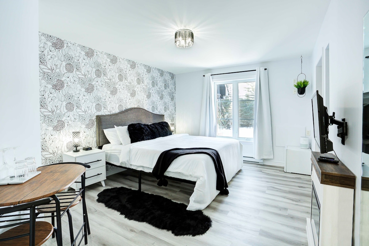 A spacious king bedroom is presented with a stylish floral accent wall. The bed is dressed in crisp white linens, complemented by a black throw. A small dining table with chairs sits nearby, and a window allows natural light to brighten the room.