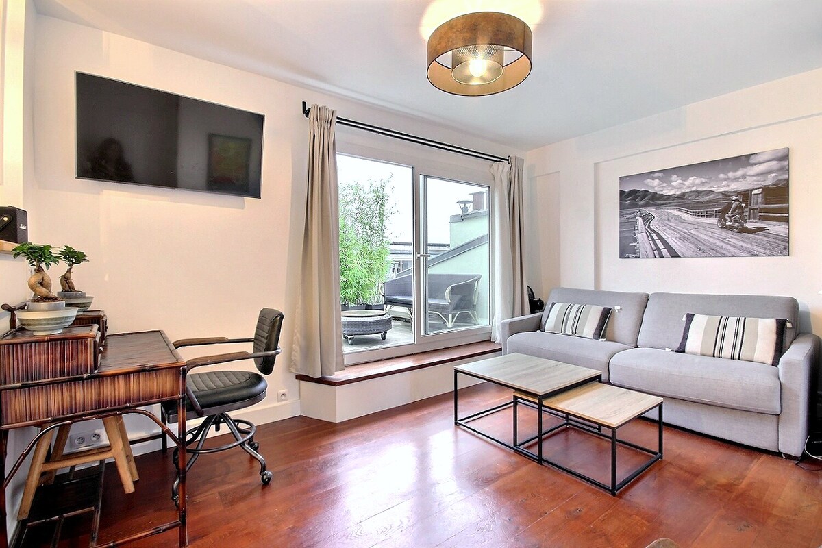 A bright living area features a comfortable gray sofa and a coffee table, complemented by a wooden desk and a chair. A large window offers views of the outdoors, while a flat-screen TV is mounted on the wall, enhancing the modern aesthetic.