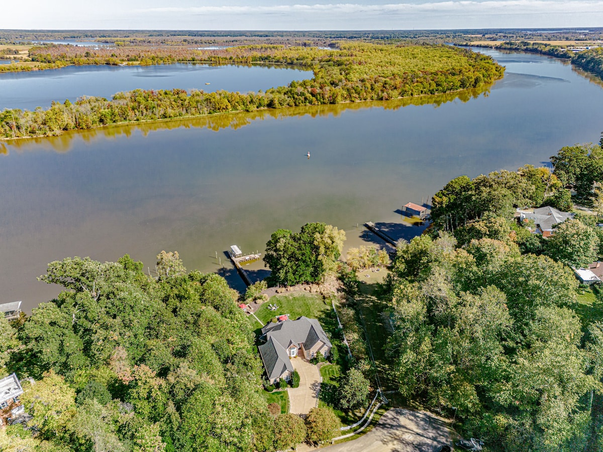 An aerial view captures the home's landscaped yard and private boat dock along the tranquil James River. Surrounding trees display vibrant autumn colors, and the serene water reflects the sky. The property is set in a peaceful, natural environment, ideal for outdoor enjoyment.