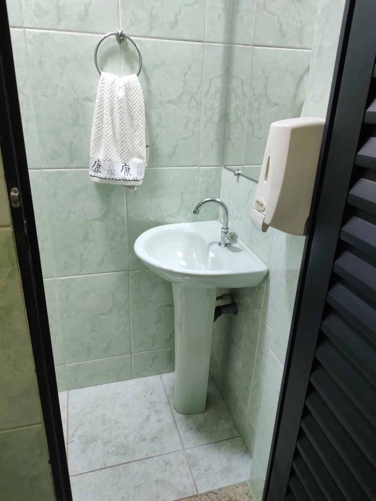 A compact bathroom is depicted, featuring light green tiled walls and a white pedestal sink. A towel is hanging on a wall rack, while a soap dispenser is mounted nearby. The floor is finished in a light-colored material, adding to the overall bright appearance.