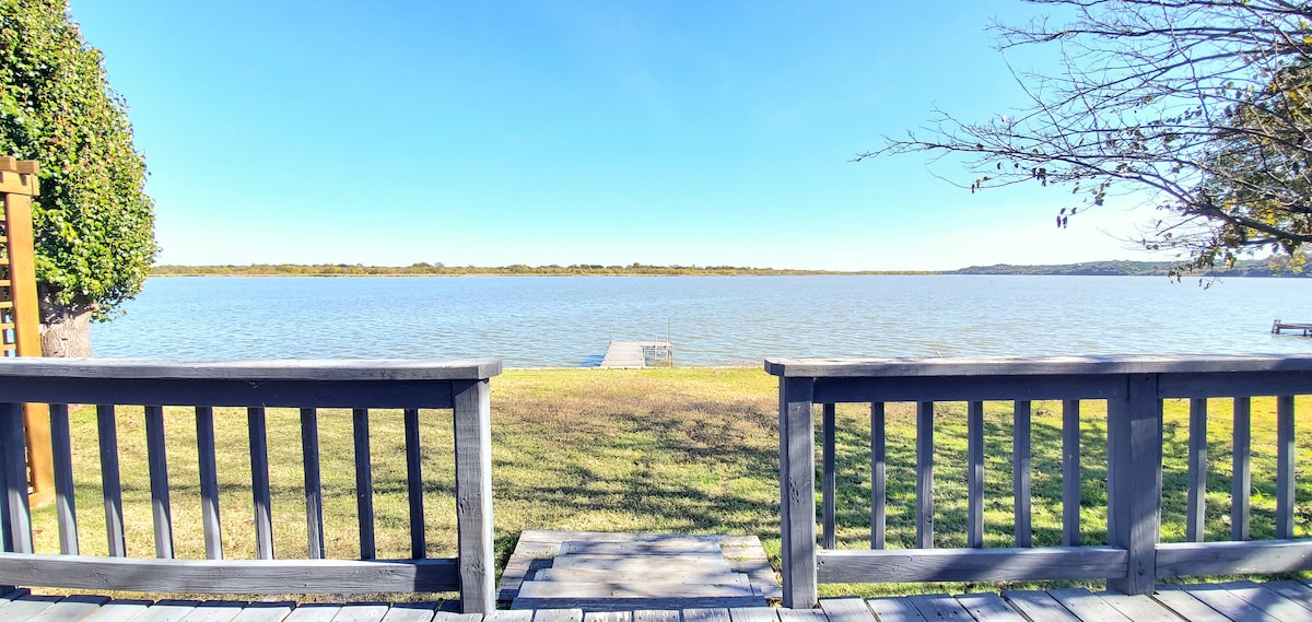 A serene view of Lake Granbury is available from the wooden deck, showcasing the water under a clear blue sky. Lush greenery borders the area, and the distant shoreline is visible, along with a dock extending into the lake.