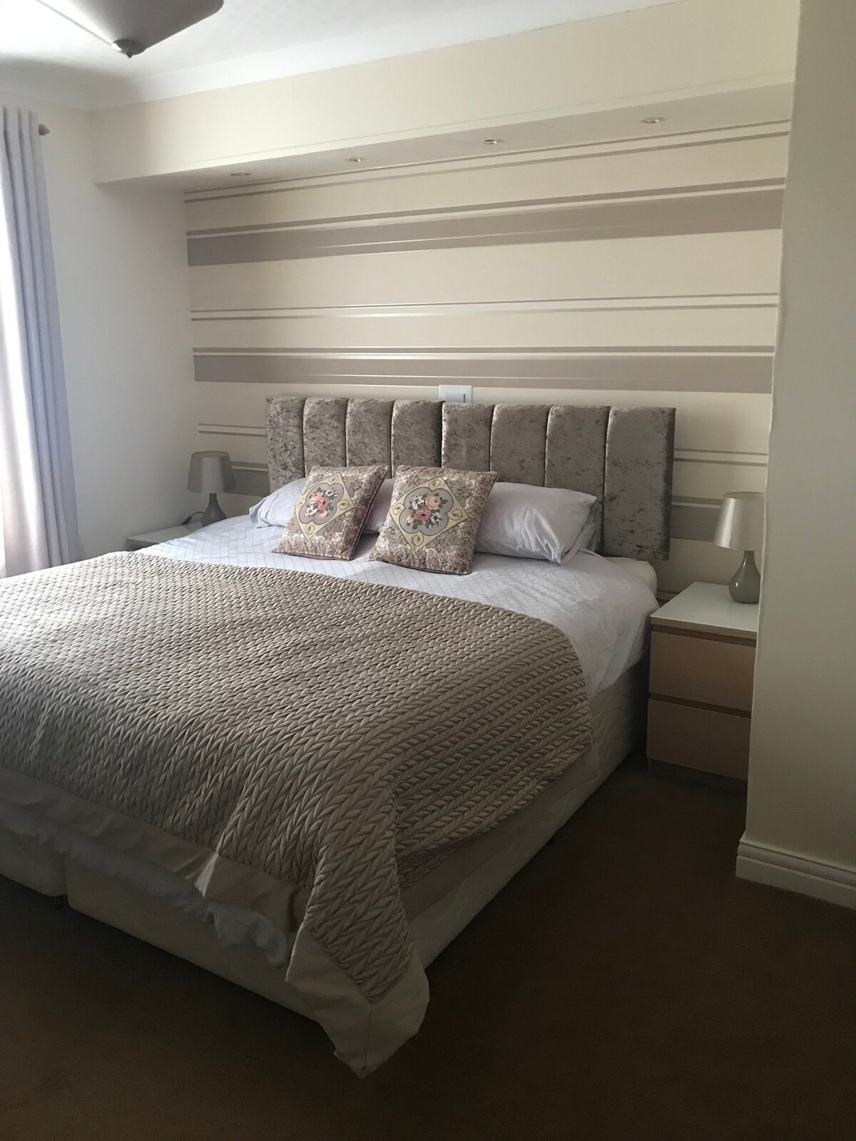 A neatly arranged bedroom features a spacious bed with a textured quilt and two patterned cushions. Nightstands are positioned on either side, each equipped with a small lamp. Natural light enters through the window, complemented by soft-colored curtains.
