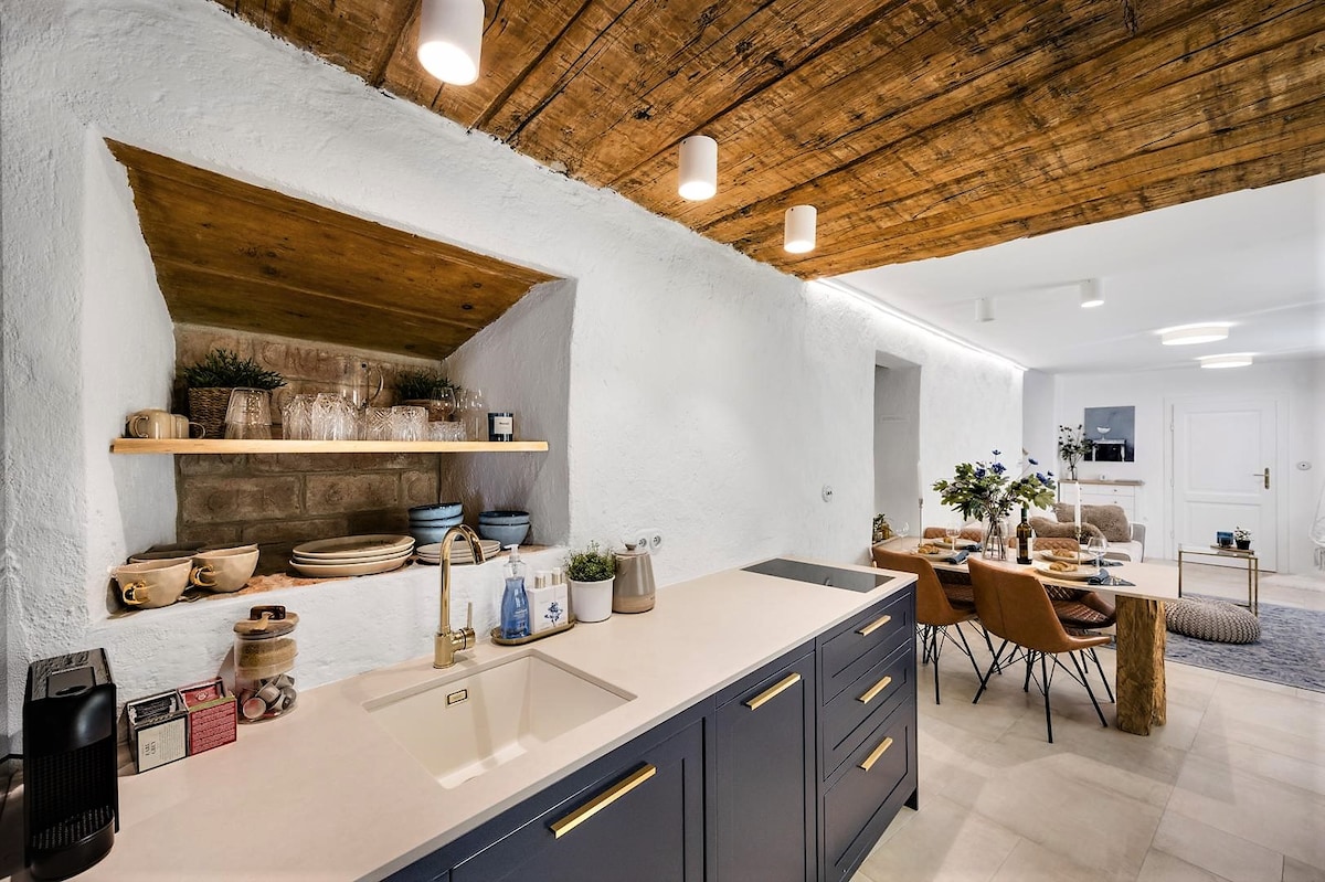 A modern kitchen area features a sleek countertop with a sink and a NESPRESSO coffee machine. Above, wooden shelving displays a variety of dishes and decorative items. The room is illuminated by ceiling lights, while the adjacent dining space is furnished with cozy brown chairs around a simple wooden table.
