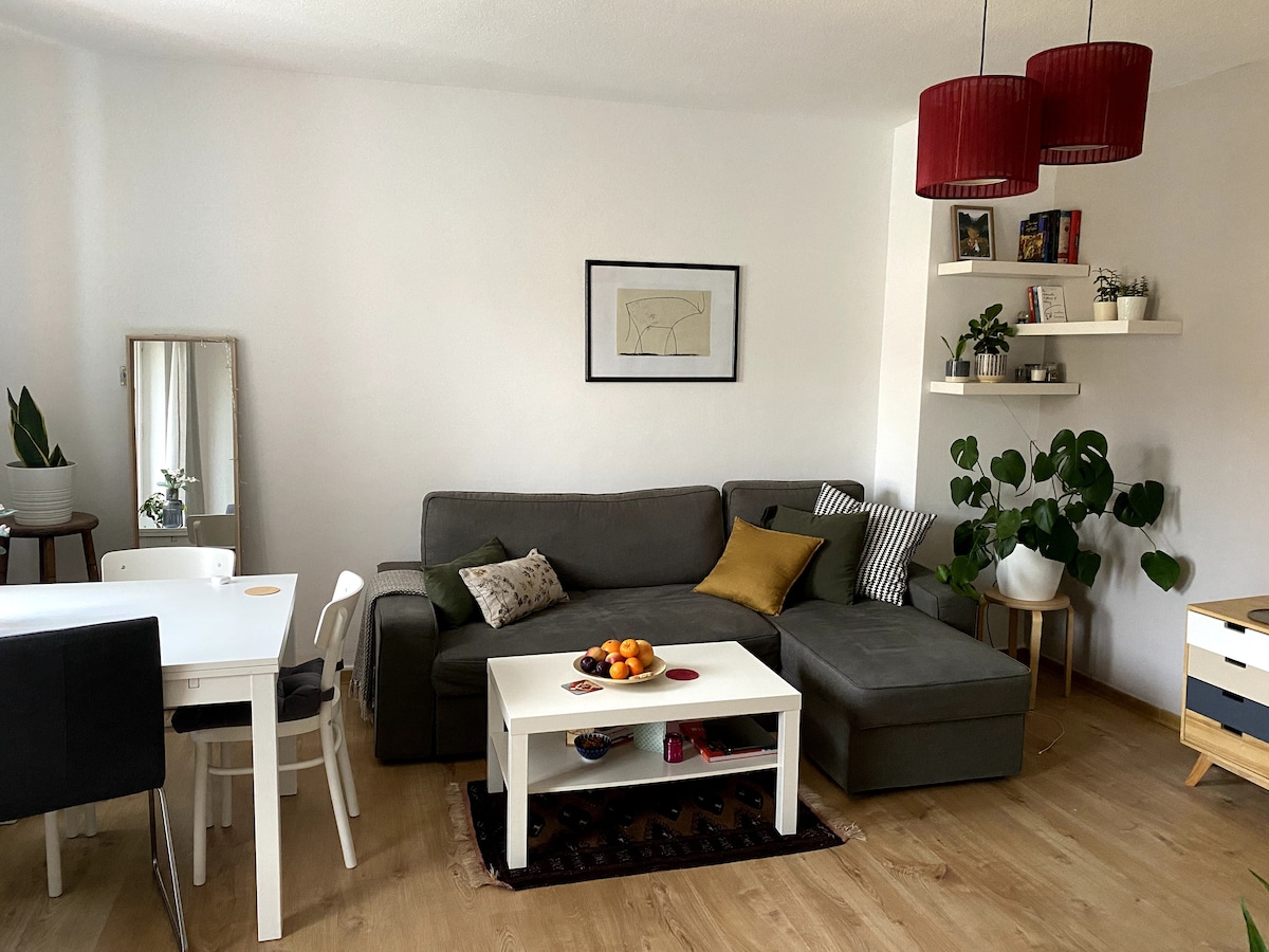 A modern living area features a comfortable gray sofa with a decorative yellow pillow. A white coffee table sits in front, adorned with a bowl of oranges. A dining table is visible, accompanied by white chairs. Green plants add a touch of nature, enhancing the space.