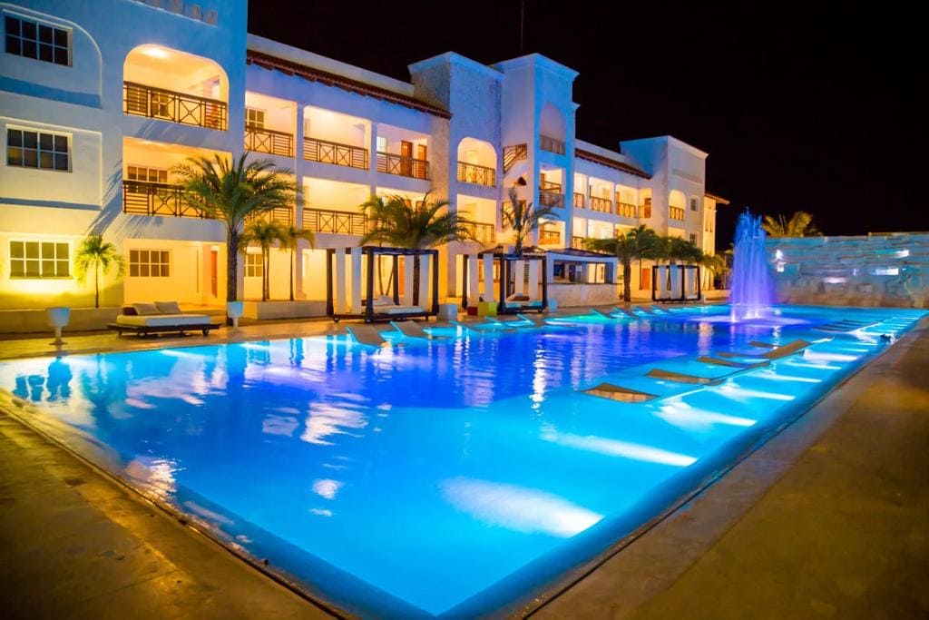 A modern resort building is illuminated at night, showcasing a large swimming pool with a serene blue hue. Lounge chairs are positioned along the pool's edge, and palm trees provide a touch of greenery in the background.