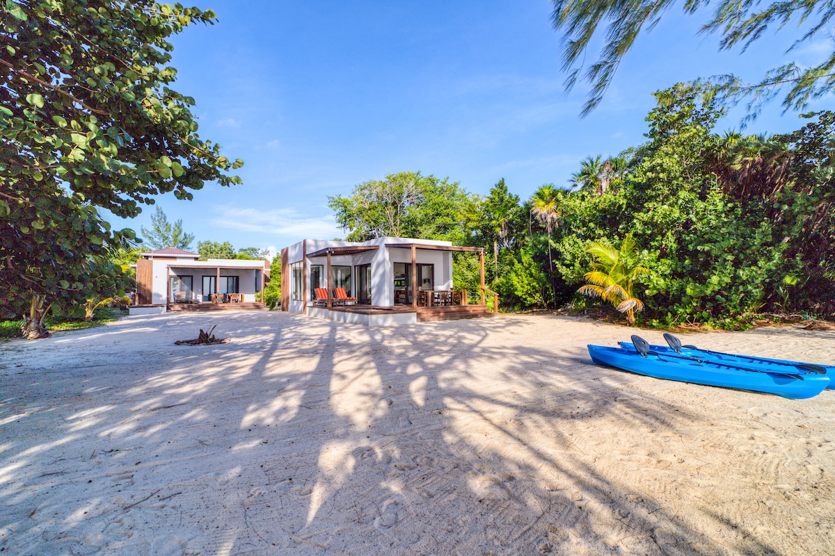 The image features a two-villa compound set on white sand, surrounded by lush coastal greenery. Two modern villas are visible, each with large windows and outdoor seating. Blue kayaks rest on the sand nearby, with shadows of trees creating a natural ambiance.