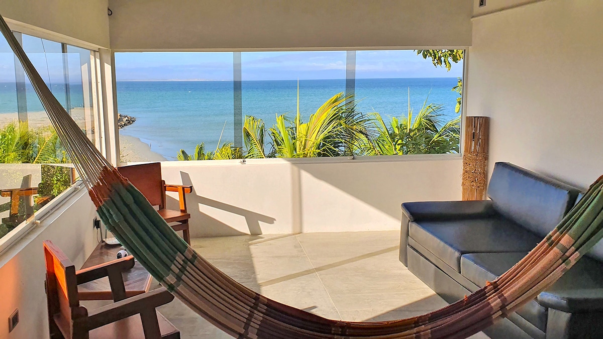 A relaxing seating area features a hammock and a sofa, positioned beside large windows that offer unobstructed views of the beach and ocean. A small table and chairs are also present, allowing for a comfortable space to enjoy the scenic surroundings.